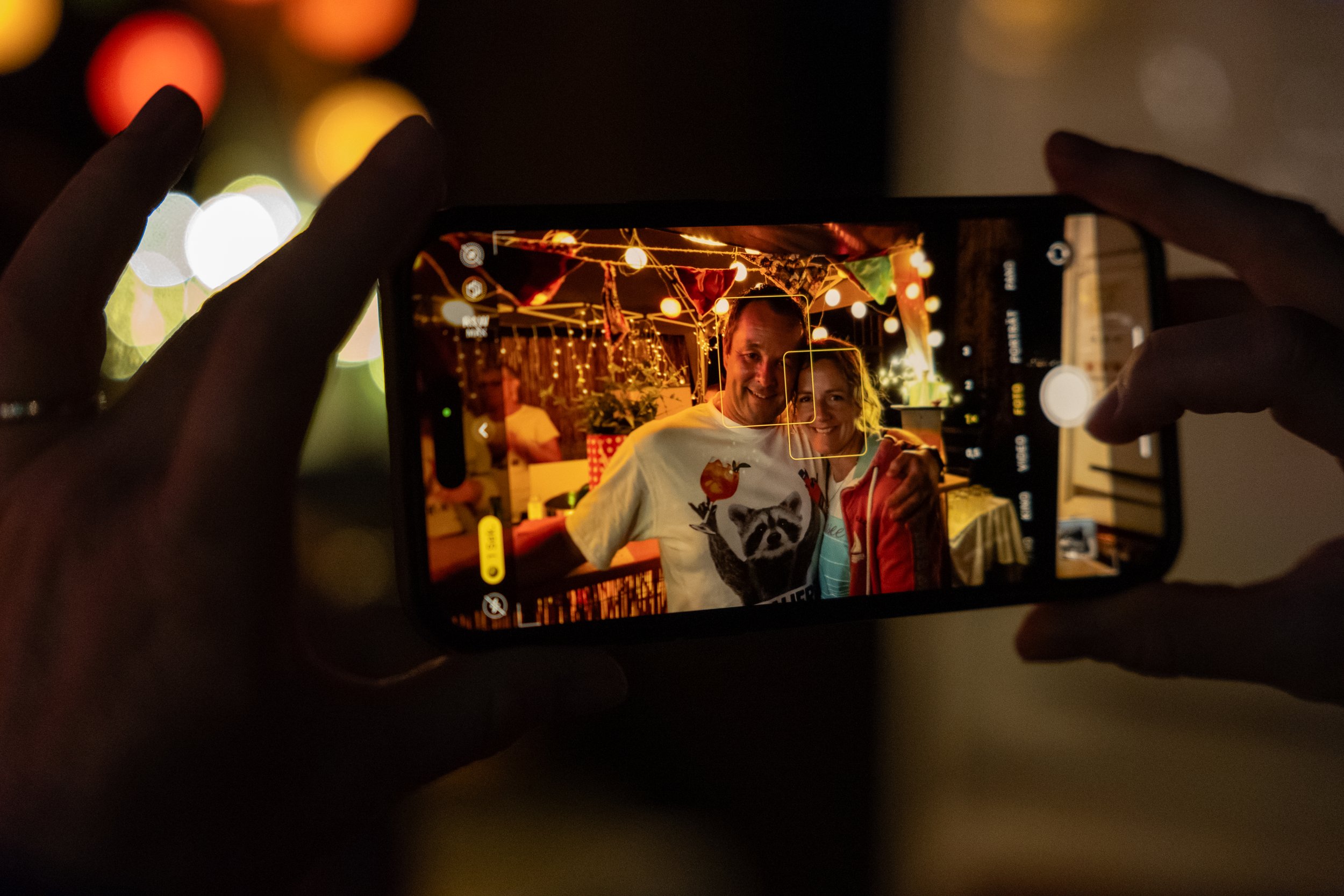 Eine Nahaufnahme von zwei Menschen, die auf einer Party oder FeierUmgebung mit Lichterketten lachen und sich umarmen, während sie ein Handy verwenden, um das Foto zu machen.