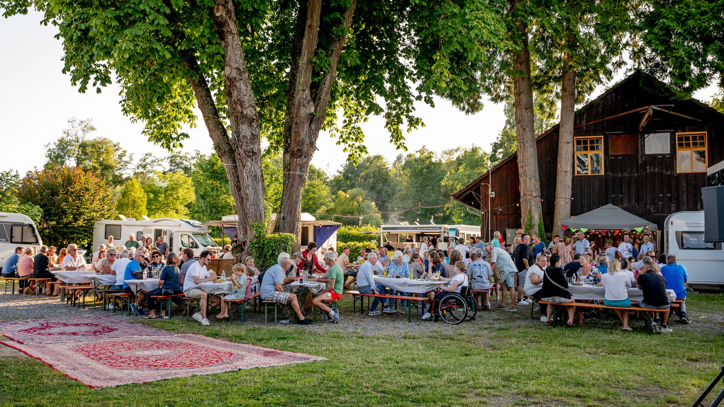 Gesellige Außengemeinschaft bei einem Picknick unter hohen Bäumen, mit Tischen, Zelten, einem alten Holzgebäude und mehreren Wohnmobilen im Hintergrund.