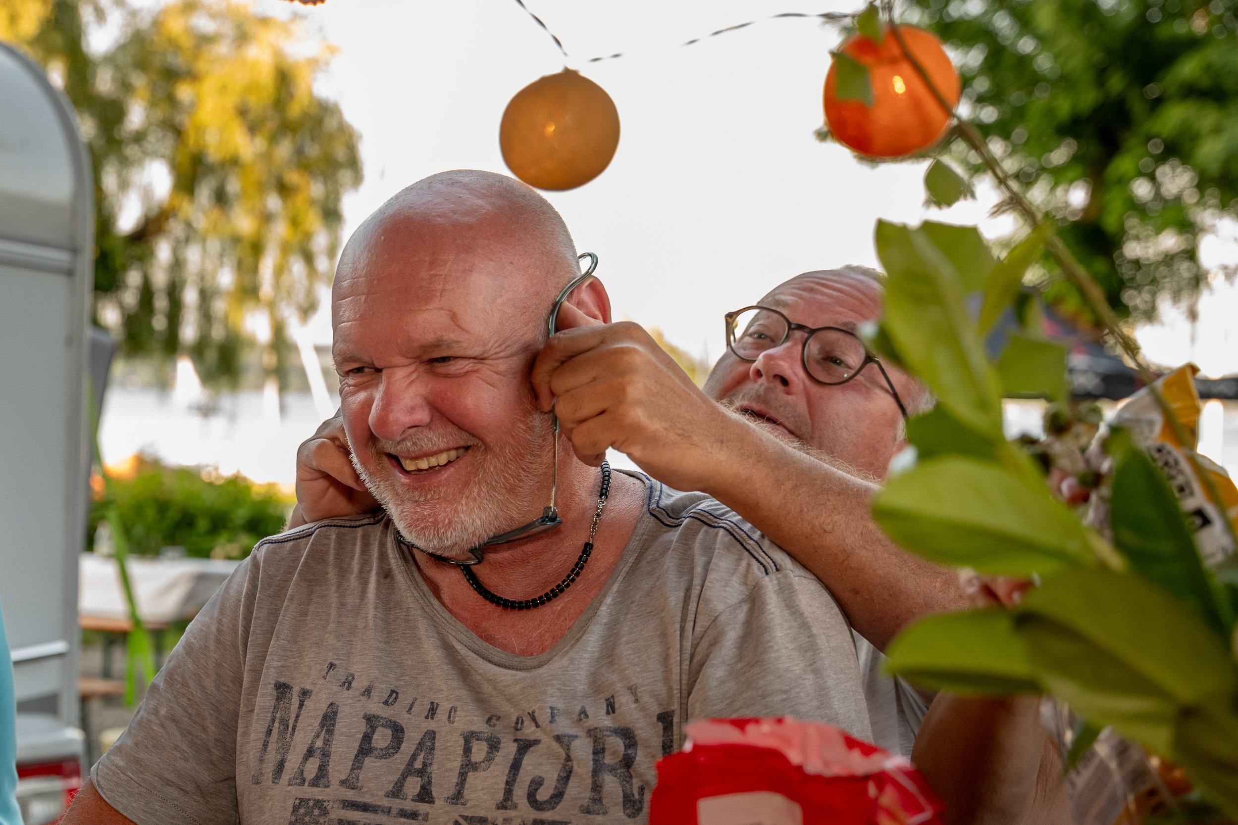 Zwei Männer, einer mit grauem Bart und Brille, beim gemeinsamen Spaß, beim Aufsetzen eines Hörers im Freien bei Sonnenuntergang, mit Obst und grünen Bäumen im Hintergrund.