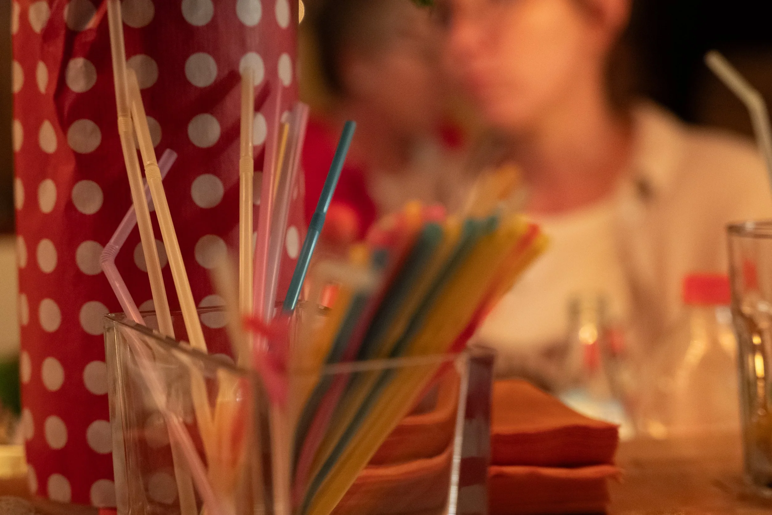 Bunte Strohhalme in einem Glas und bunte Papierstreifen auf einem Tisch, im Hintergrund eine Frau mit einem weißen Oberteil, unscharf.
