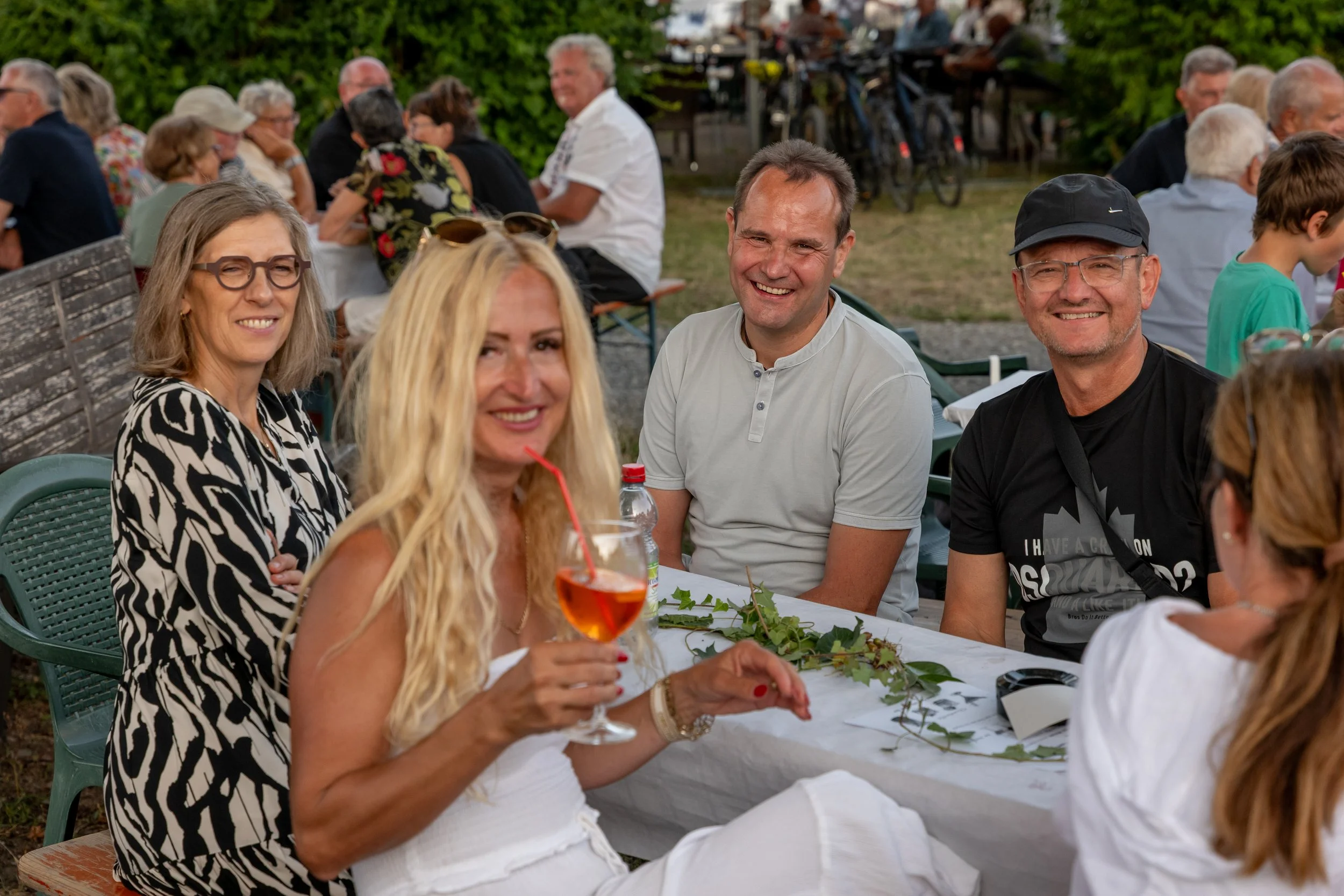 Gruppe von Menschen sitzt an einem festlichen Tisch im Freien, einige lachen, eine Frau hält ein Getränk mit einem roten Strohhalm, es gibt Getränke und Dekorationen auf dem Tisch, im Hintergrund andere Gäste und Bäume.