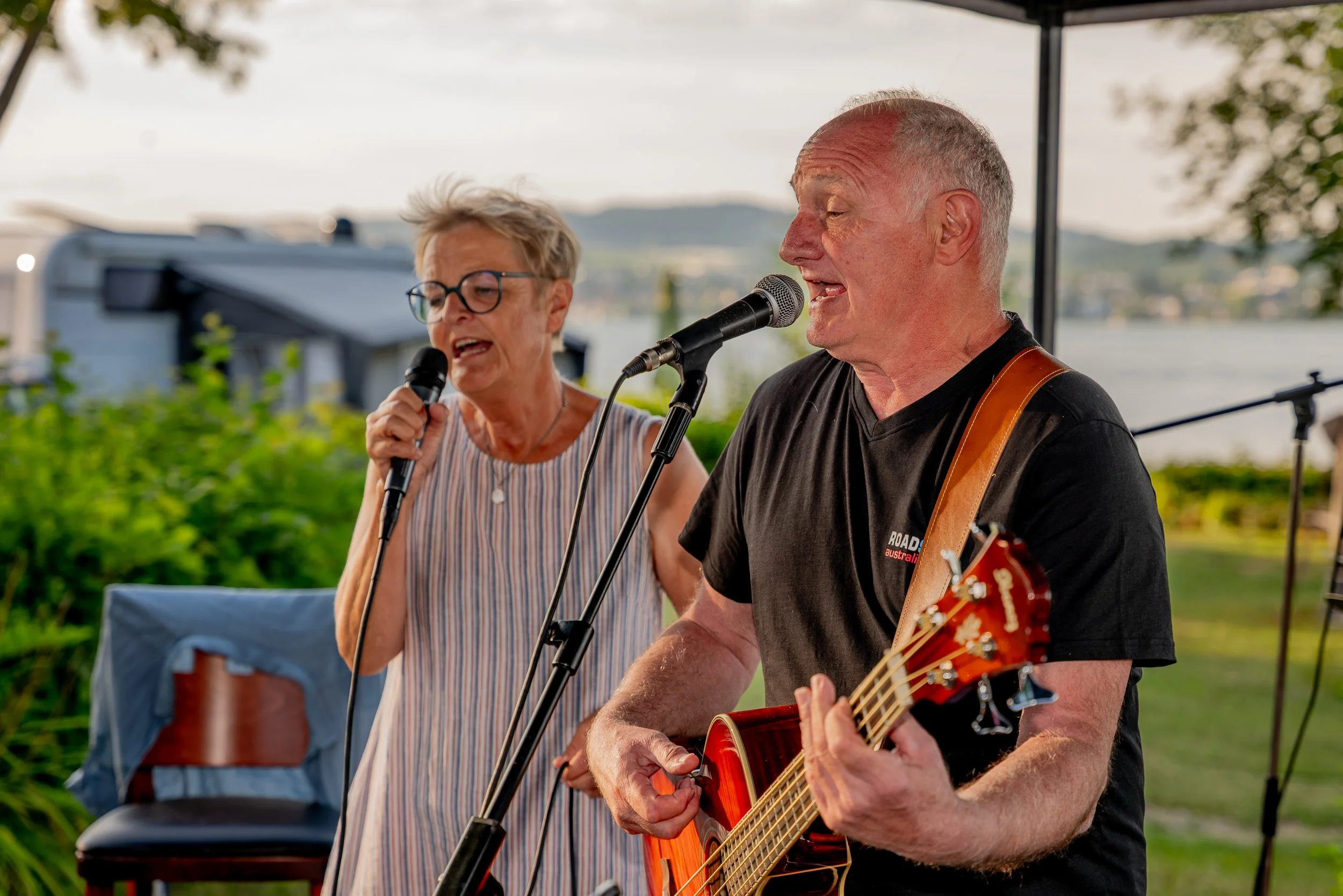 Ein älterer Mann spielt Gitarre und singt, während eine ältere Frau neben ihm singt, beide im Freien bei Sonnenuntergang, mit Wasser im Hintergrund.
