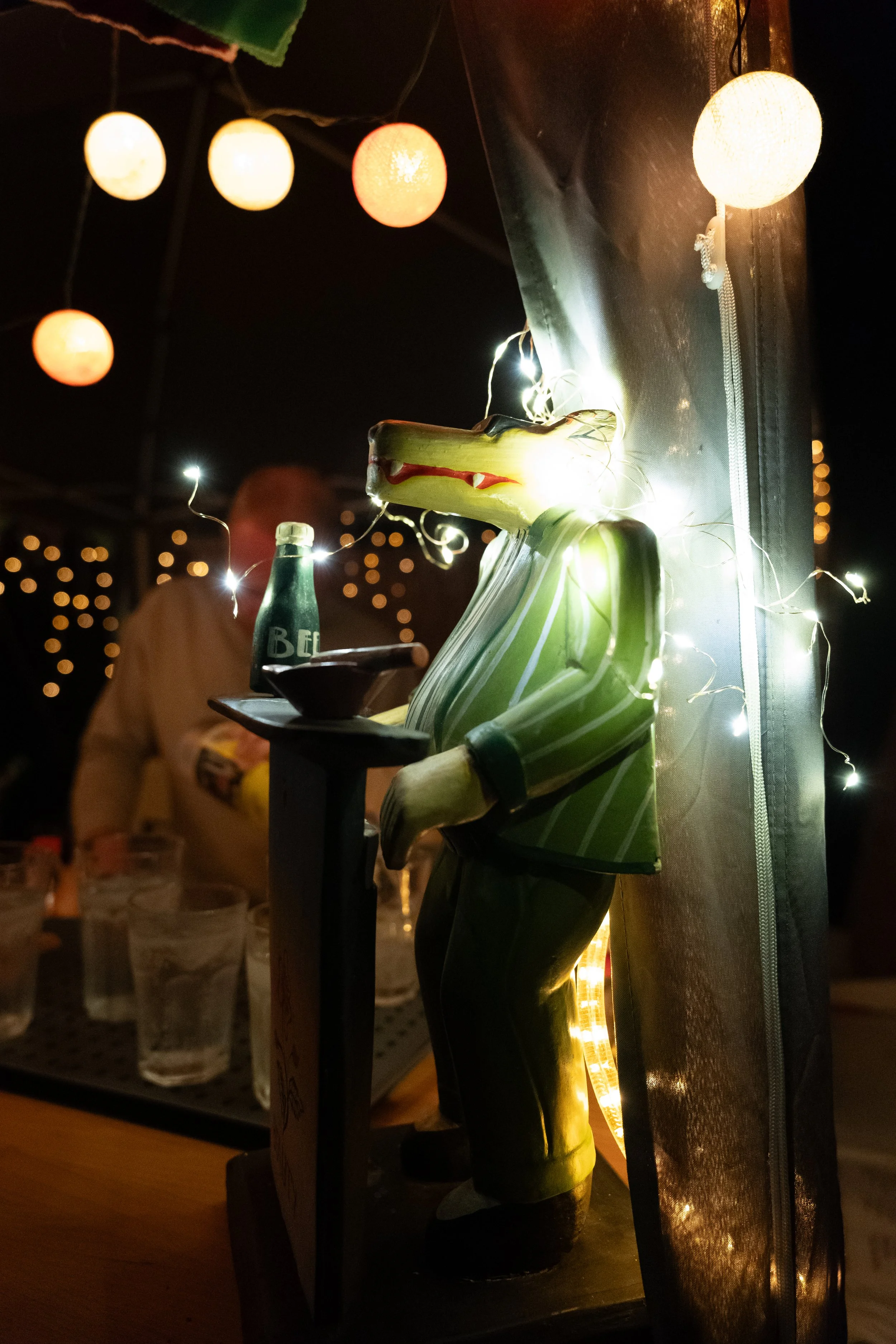 Eine Krokodil-Figur in einem Restaurant bei Nacht, die einen Anzug trägt und eine Flasche sowie eine Schale hält. Im Hintergrund sind Menschen und bunte Lichter zu sehen.