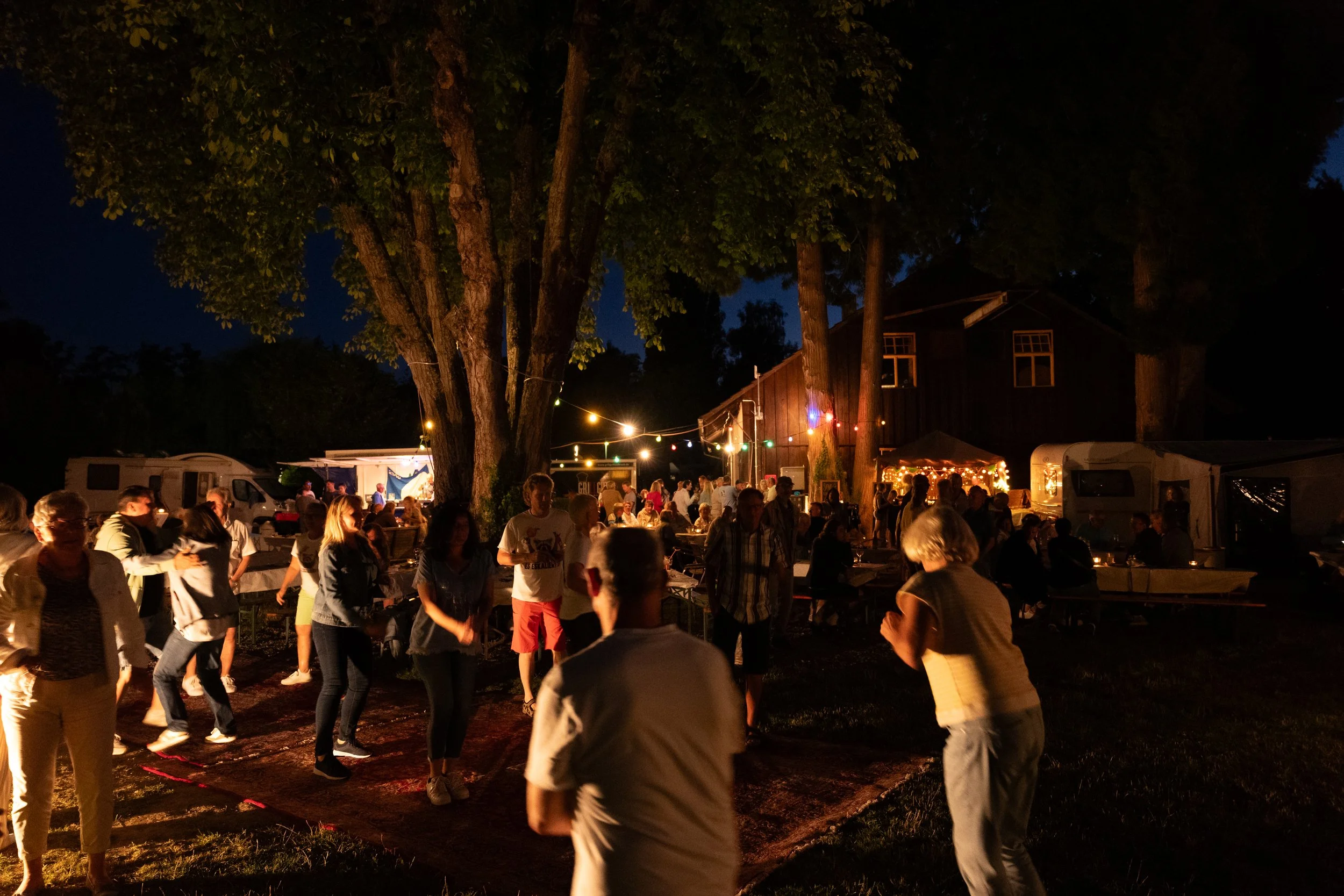 Menschen tanzen bei einer nächtlichen Feier im Freien unter Lichterketten neben einem großen Baum, umgeben von Zelten und einer Holzhütte.
