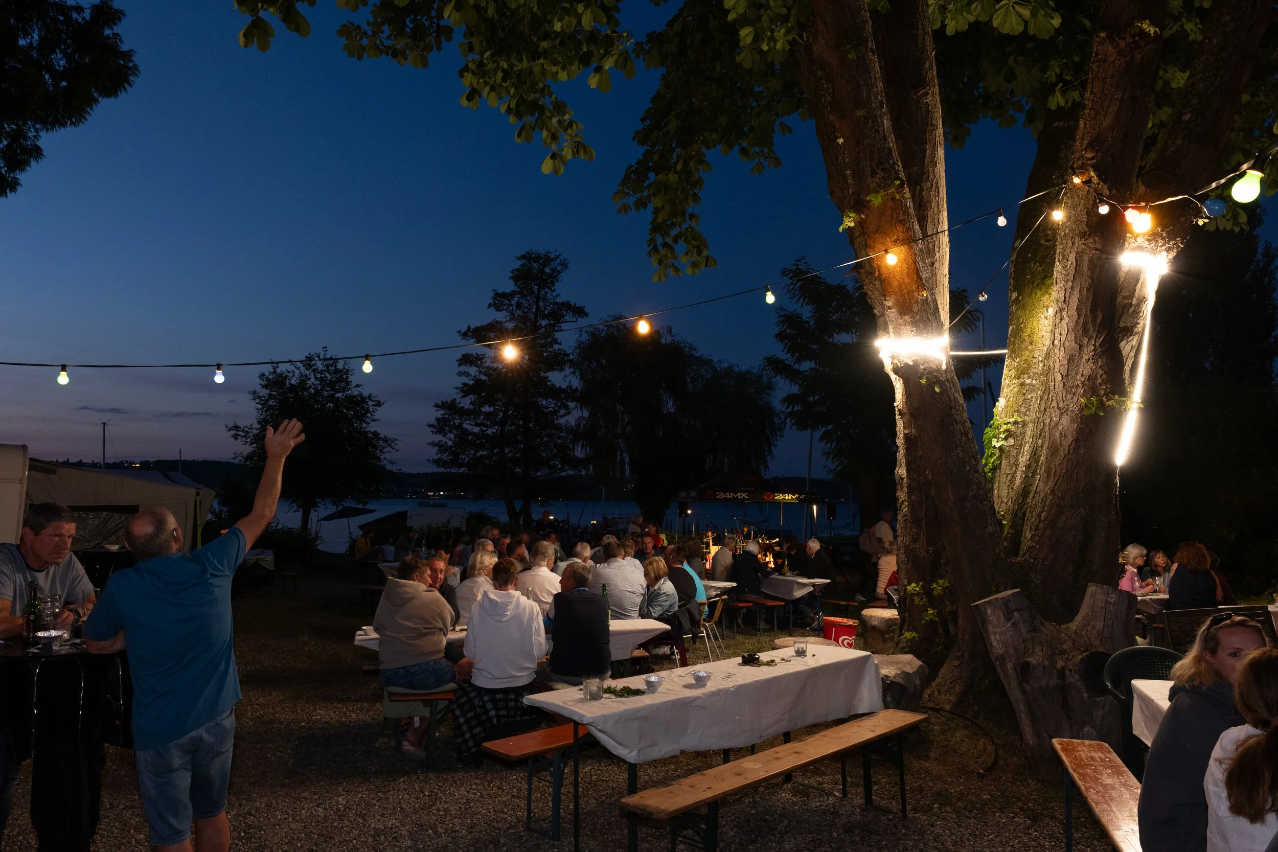 Abendliches Treffen im Freien mit mehreren Menschen, Tischen und Lichterketten, umgeben von Bäumen am Wasser, bei Sonnenuntergang