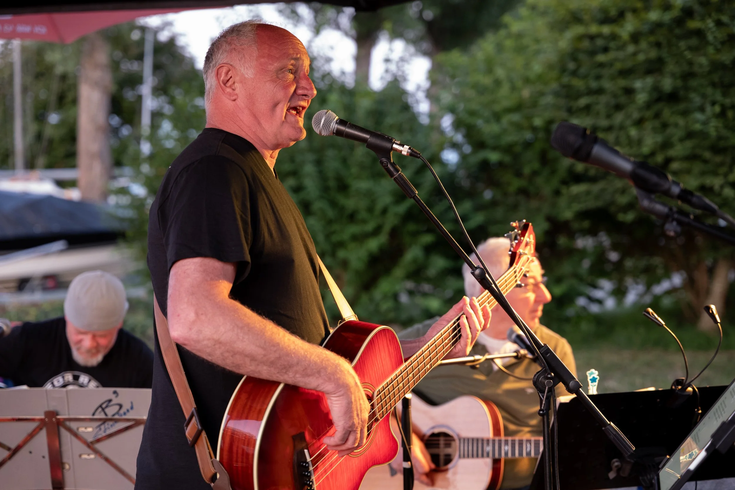 Eine Gruppe von Musikern spielt Gitarre und singt im Freien bei Sonnenuntergang, umgeben von grünen Bäumen.