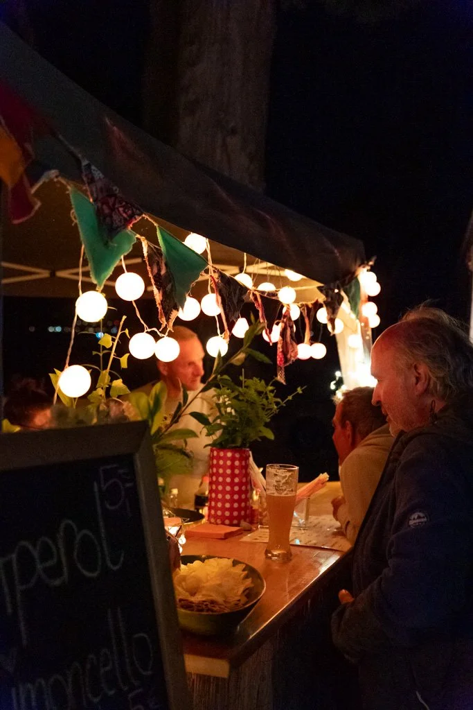 Menschen sitzen an einem mit Lichterketten dekorierten Stehtisch bei einem nächtlichen Event im Freien.