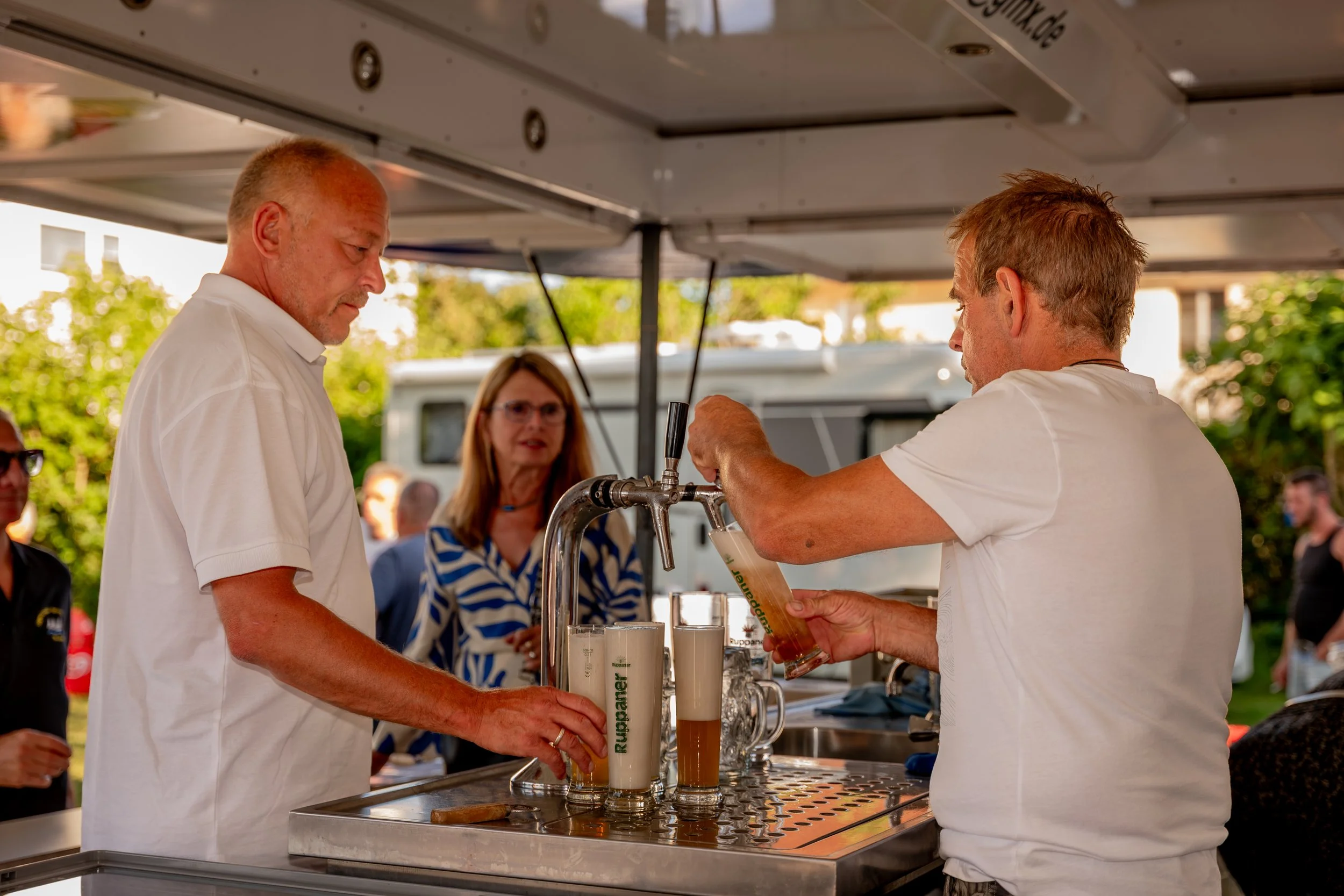Ein Bierwagen, an dem zwei Männer Bier einschenken, während eine Frau im Hintergrund zuschaut. Es ist Sonnenwetter im Freien mit grünen Bäumen im Hintergrund.