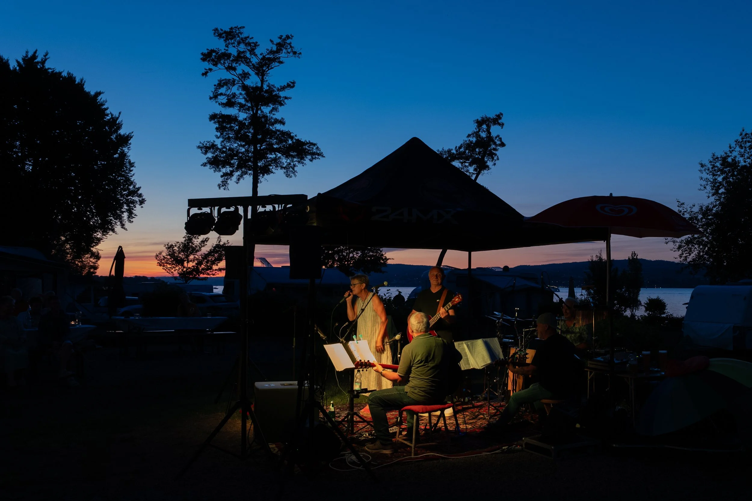 Musikgruppe spielt bei Sonnenuntergang im Freien unter einem Zelt am Wasser, umgeben von Bäumen und Campingzelten.