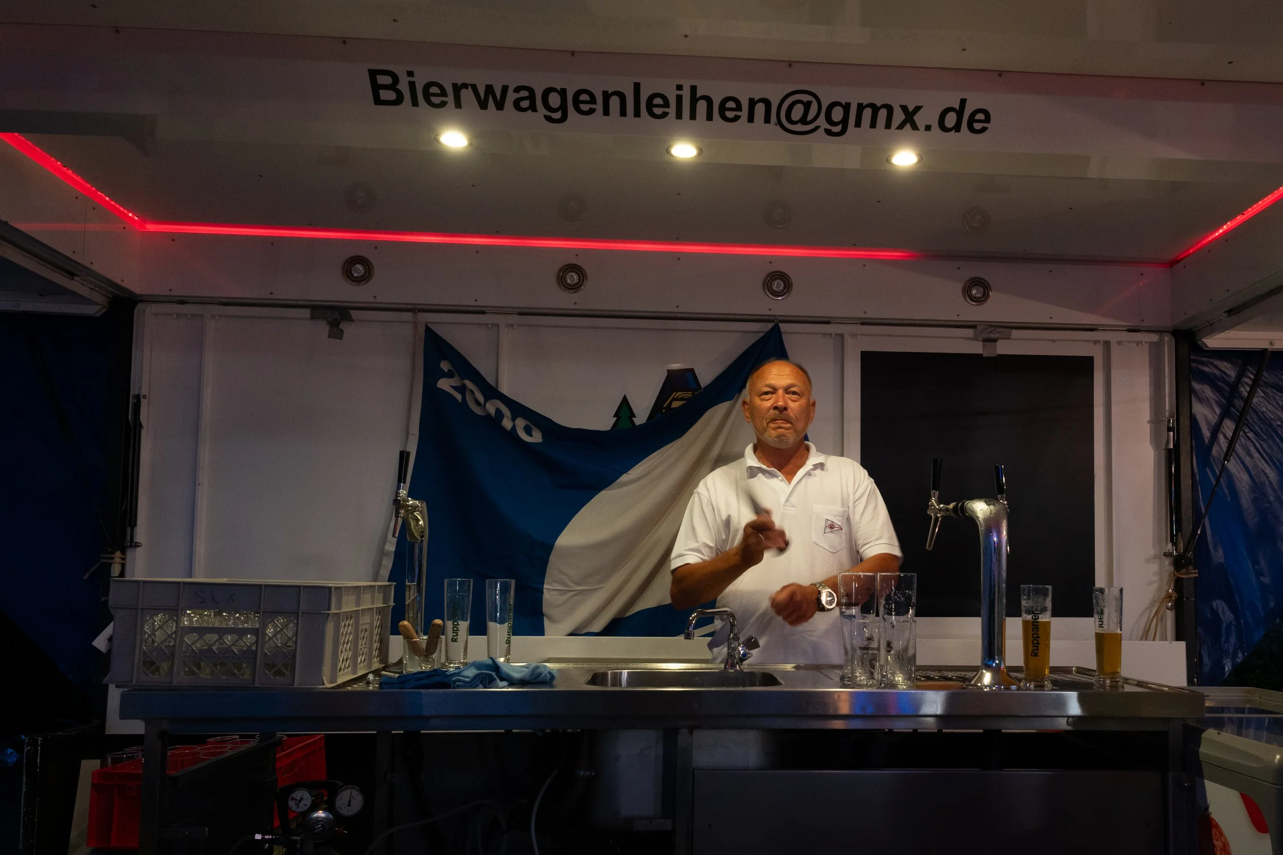 Mann hinter einem Bar-Stand mit Bier-Gläsern, hinter ihm Fahne und ein Banner mit der E-Mail-Adresse "Bierwagenleihen@gmx.de"