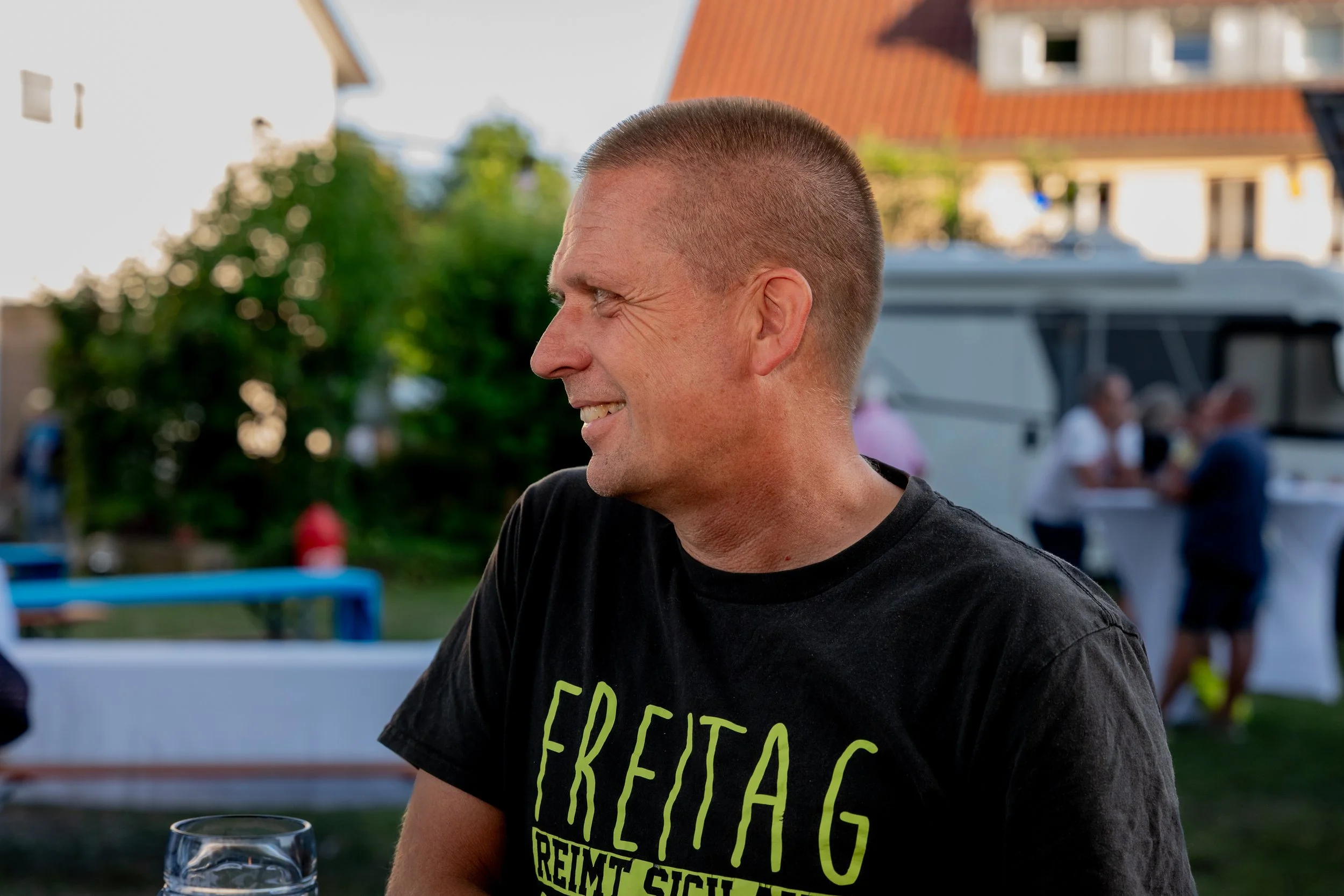 Ein Mann in einem schwarzen T-Shirt, der im Freien beim Sommerfest sitzt, lächelt und hält ein Glas. Im Hintergrund sind Bäume, Häuser und andere Menschen auf einer Feier sichtbar.