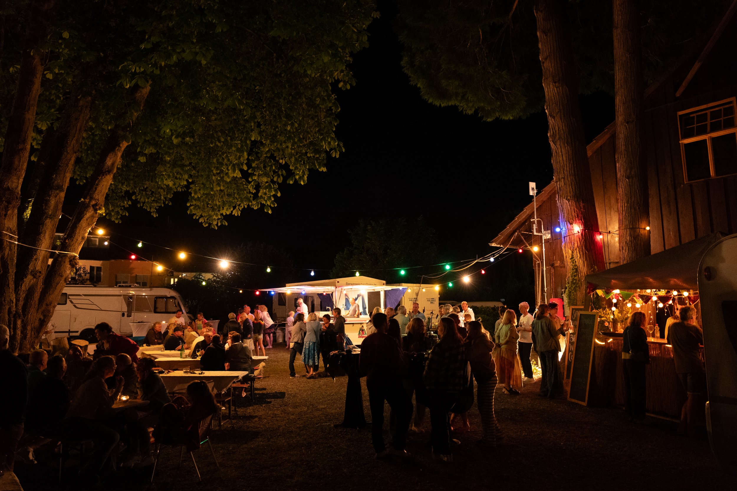 Abendliche Außenaufnahme eines Festes mit bunten Lichterketten, Menschen beim Essen und Trinken, einer Bar und einem Bühnenauftritt unter Bäumen, bei Nacht.
