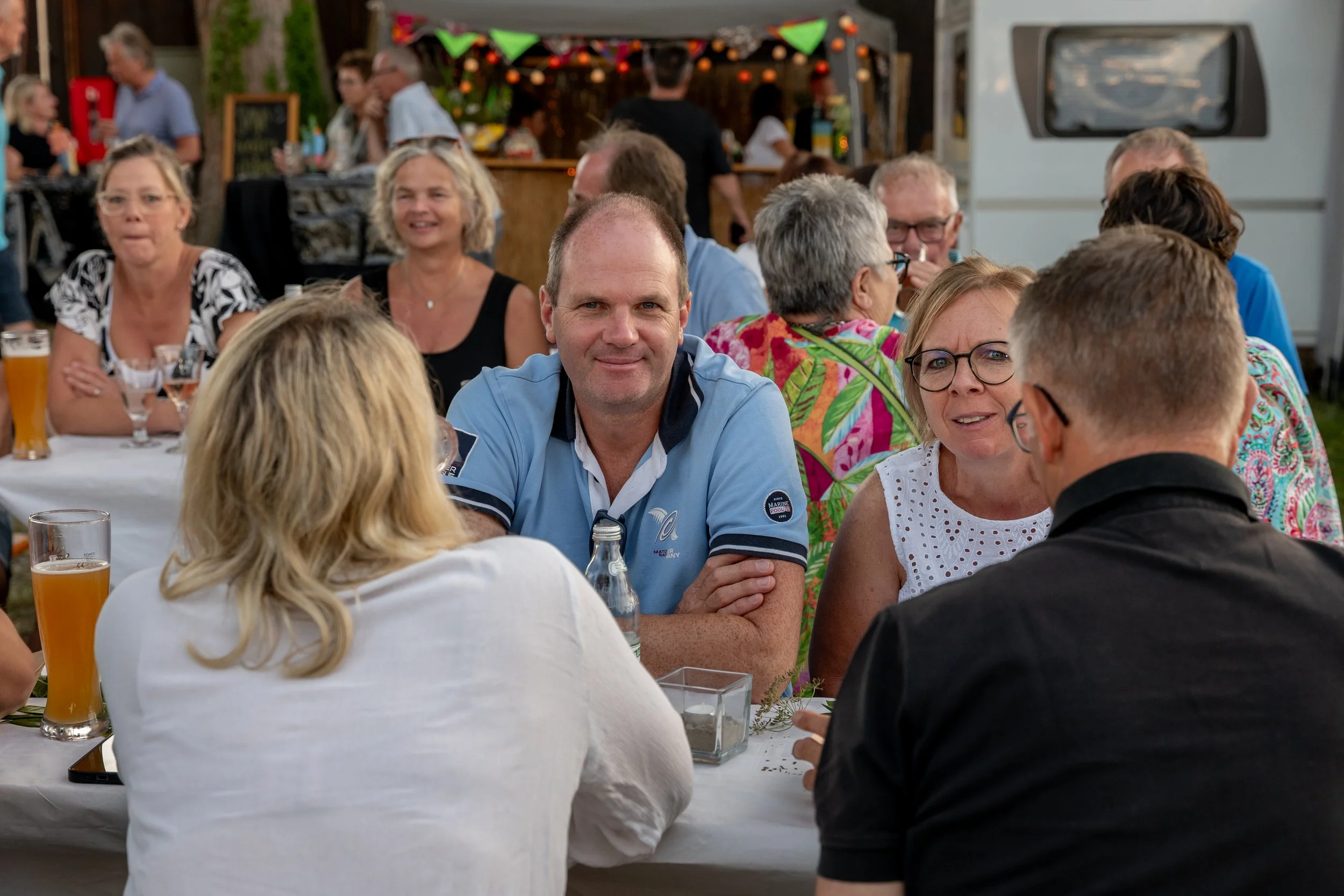Menschen sitzen bei einem Fest im Freien an einem langen Tisch, trinken und unterhalten sich, im Hintergrund dekorative Lichter und ein food-Stand.