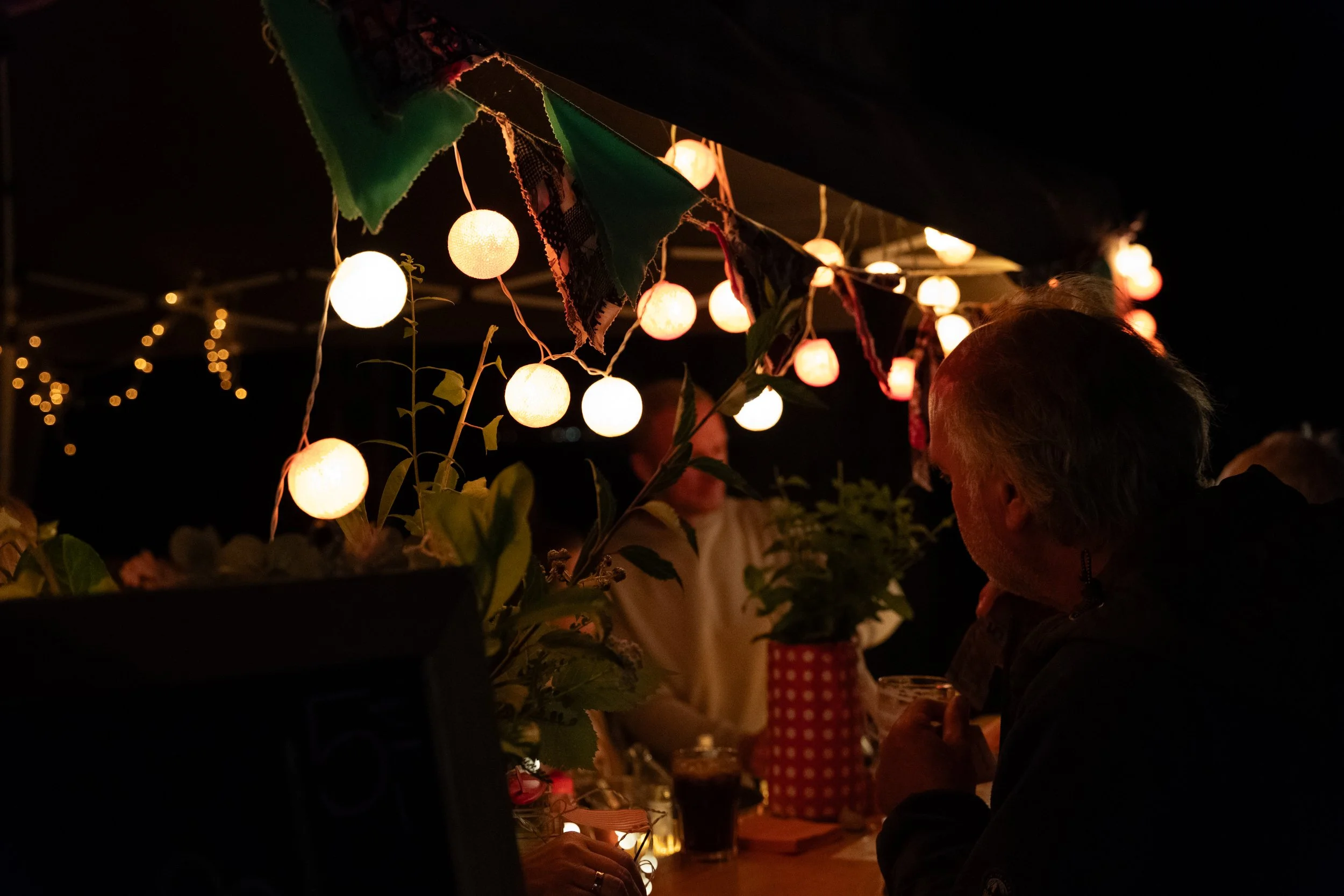 Menschen sitzen an einem Tisch unter einer Himmel voller Lichterketten und Papierlaternen, bei Nacht, vermutlich auf einem Markt oder Festival, mit dunklem Himmel im Hintergrund.