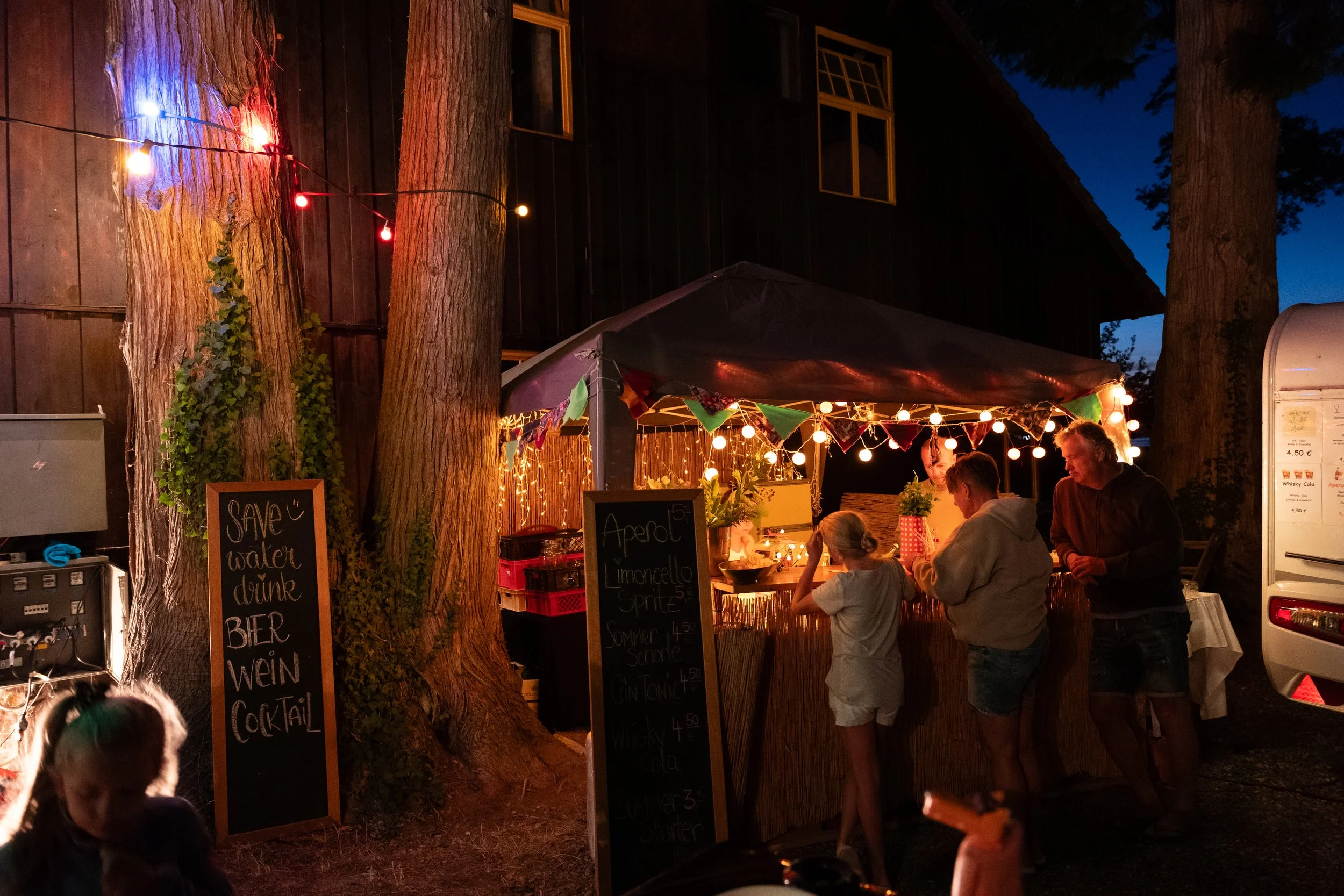 Abendliche Szene an einem kleinen Feststand mit bunten Lichterketten, einer Tafel mit Getränkekarten, mehreren Personen, die Getränke bestellen, und zwei großen Bäumen im Hintergrund.