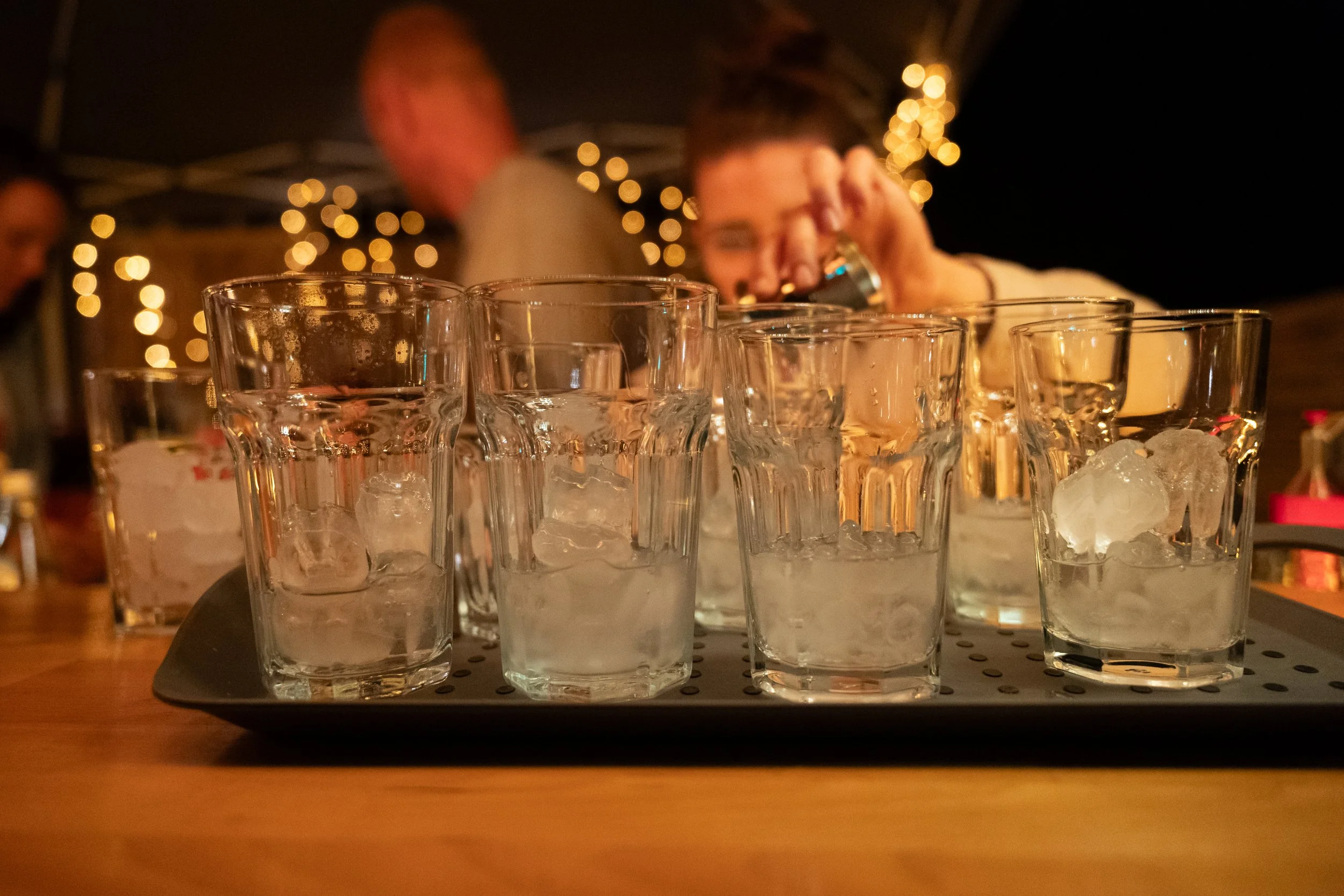 Mehrere Gläser mit Eiswürfeln auf einem Tablett, im Hintergrund eine Person, die einen Shot trinkt, bei stimmungsvoller Beleuchtung mit Lichterketten.