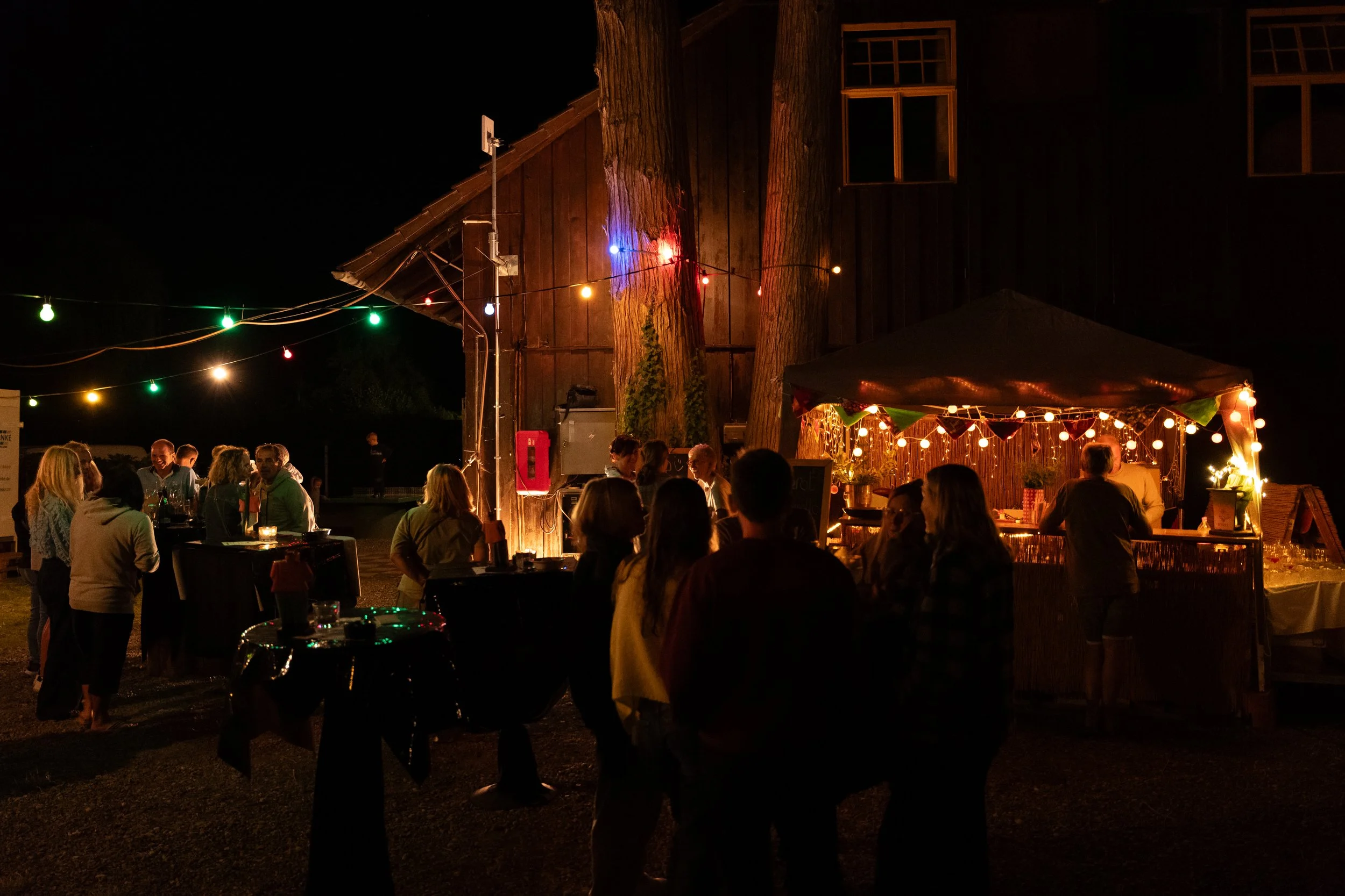 Menschen feiern nachts im Freien mit Lichterketten und einem Pavillon, der mit Lichtergirlanden dekoriert ist, vor einer Holzhütte.