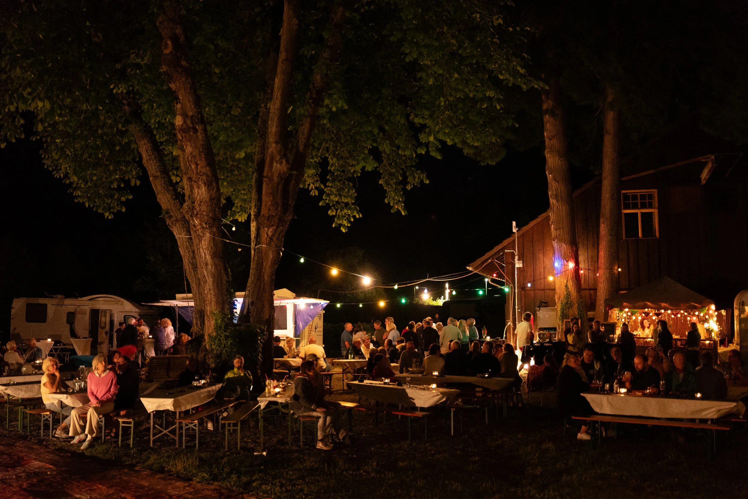Menschen feiern bei Nacht im Freien mit Lichterketten, Tischen und einem Holzgebäude im Hintergrund.