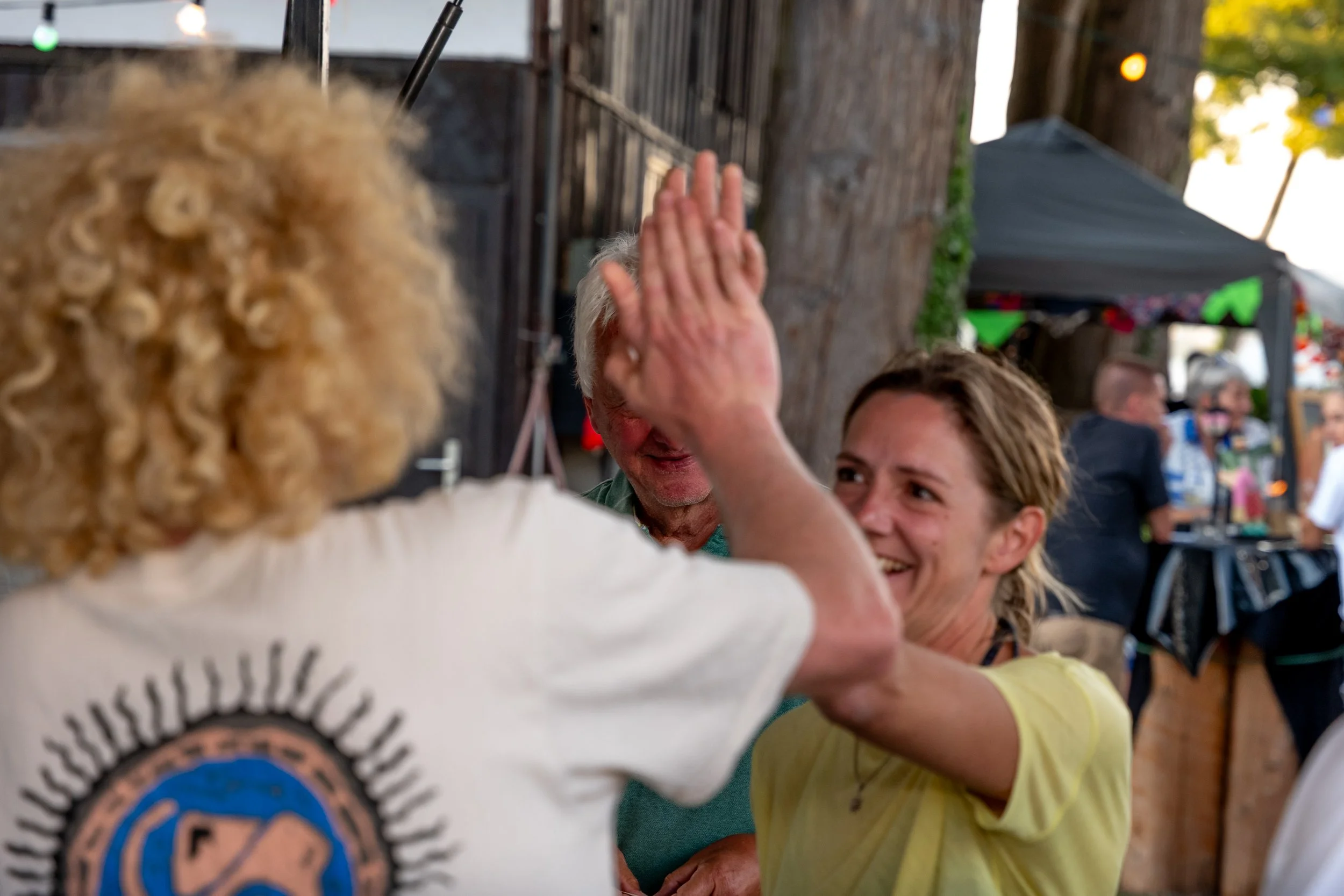 Menschen beim High Five bei einem Treffen im Freien, unter Bäumen mit einer Zelt- und Festzeltdekoration im Hintergrund.