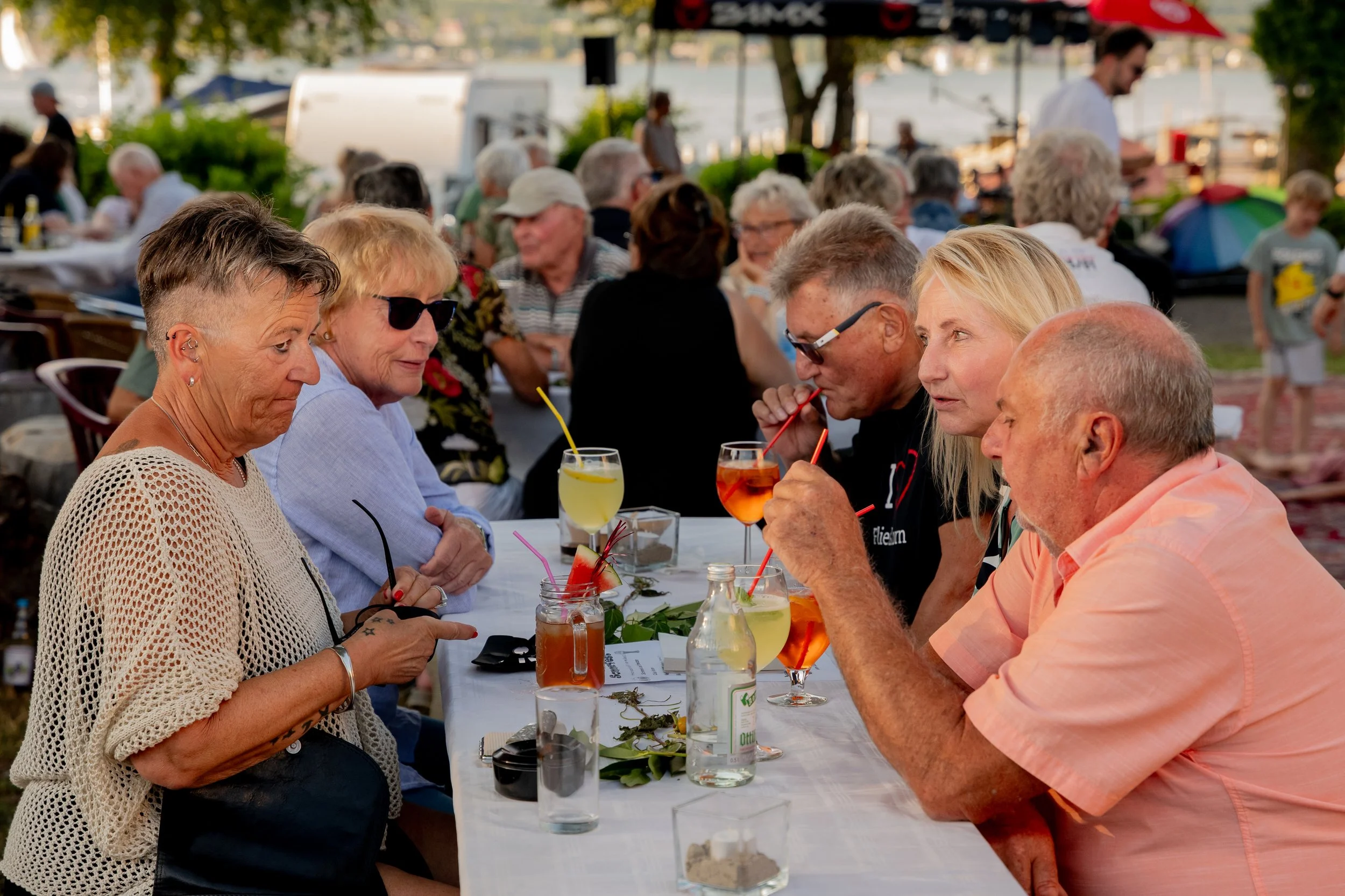 Menschen sitzen bei einem Sommerfest an einem langen Tisch im Freien, trinken cocktails und genießen die Gesellschaft, im Hintergrund sind weitere Gäste und bunte Sonnenschirme zu sehen.