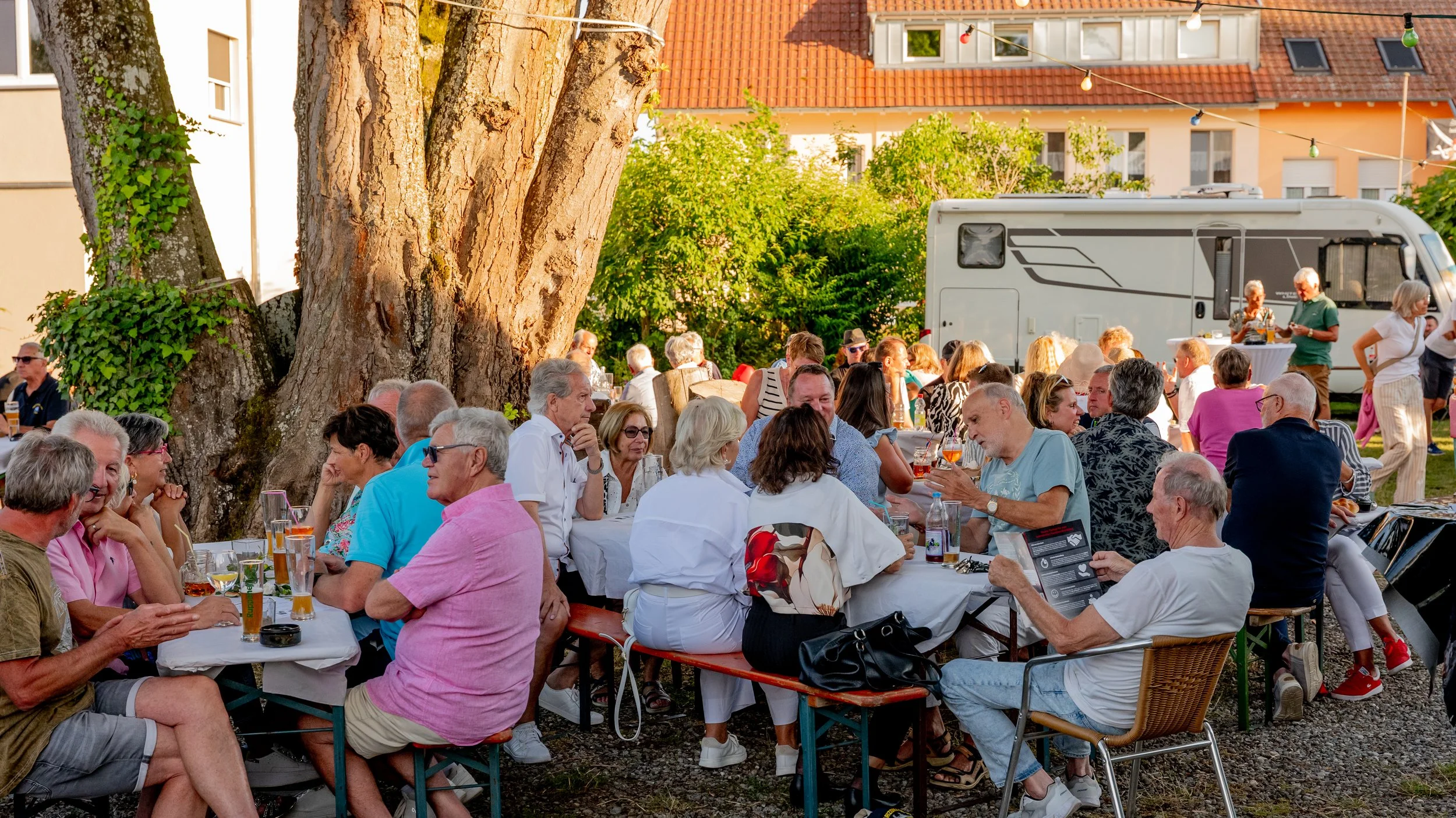 Menschen sitzen bei einem Treffen im Freien unter großen Bäumen an Tischen, es gibt Getränke und Essen, im Hintergrund ist ein Wohnmobil und mit Lichterketten geschmückt.