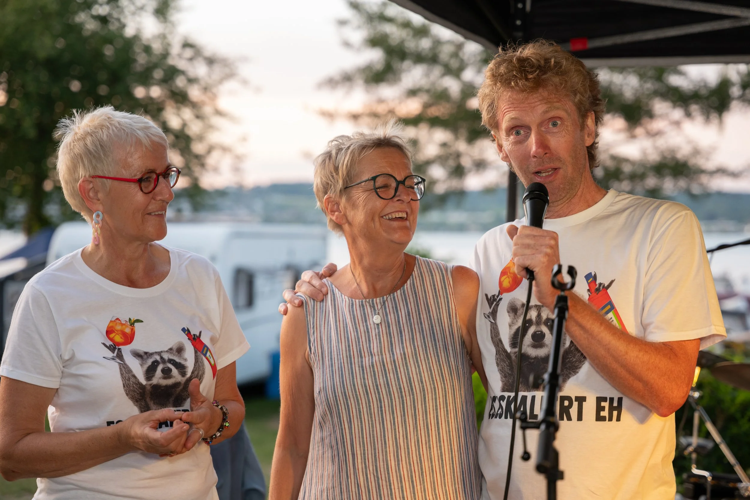 Drei Menschen stehen zusammen bei einer Veranstaltung am Wasser. Der Mann spricht in ein Mikrofon, zwei Frauen lachen und schauen ihn an. Es ist Abend oder Sonnenuntergang im Hintergrund.