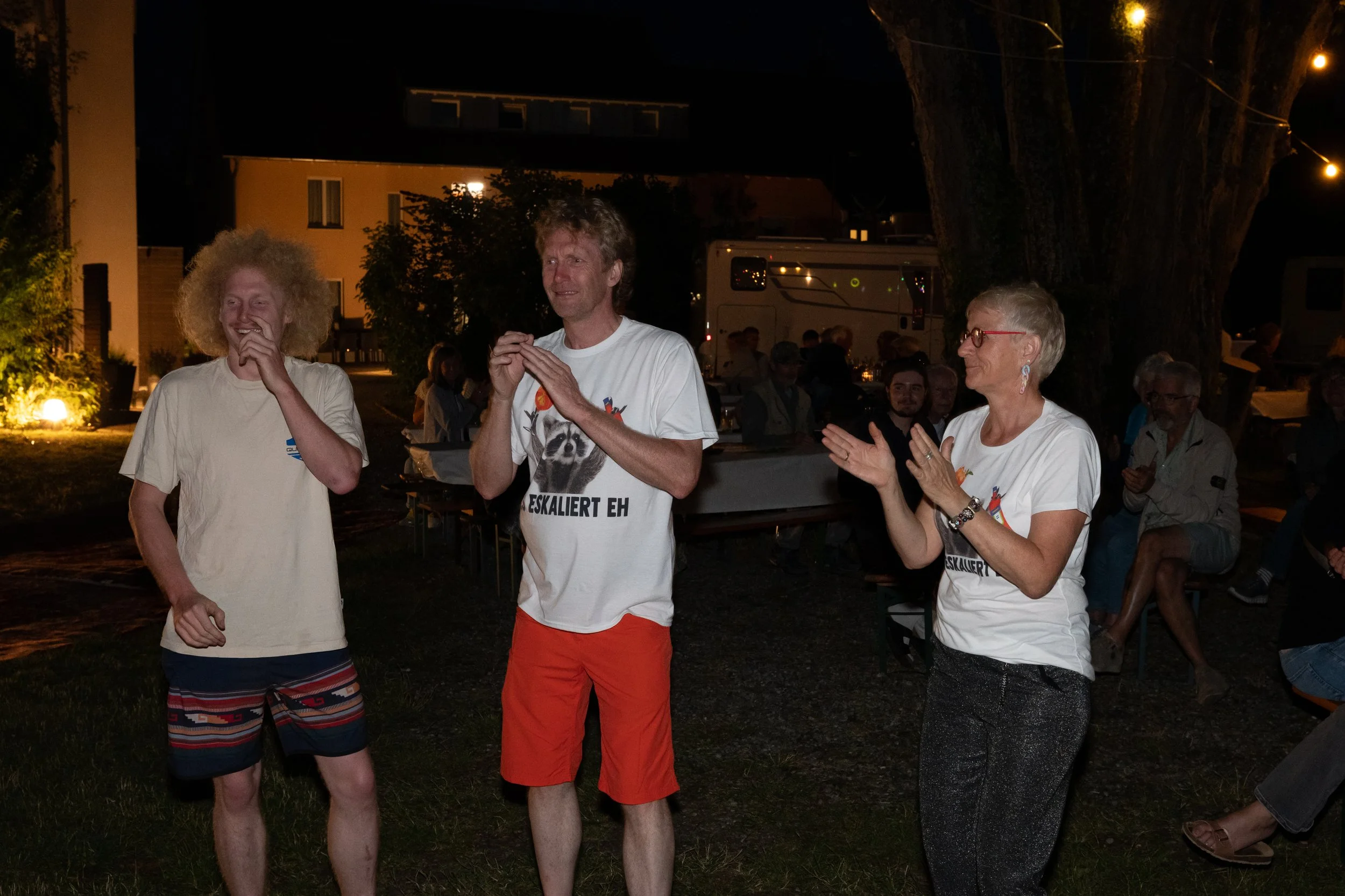Drei Menschen stehen im Freien bei Nacht, lachen und klatschen, während eine Gruppe von Zuschauern im Hintergrund sitzt.
