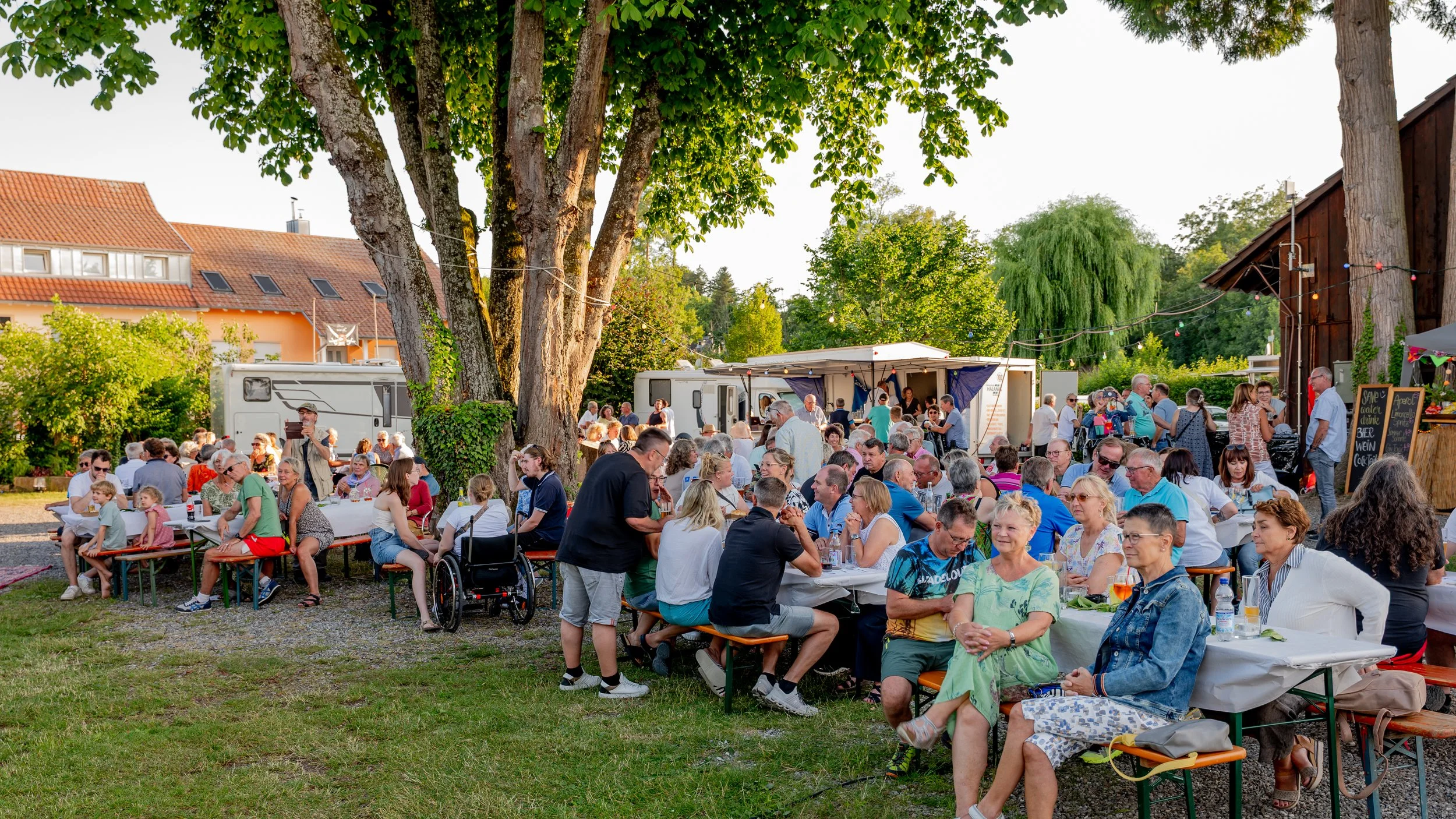 Menschen sitzen bei einem Sommerfest im Freien an Tischen, mit Bäumen und Zelten im Hintergrund.