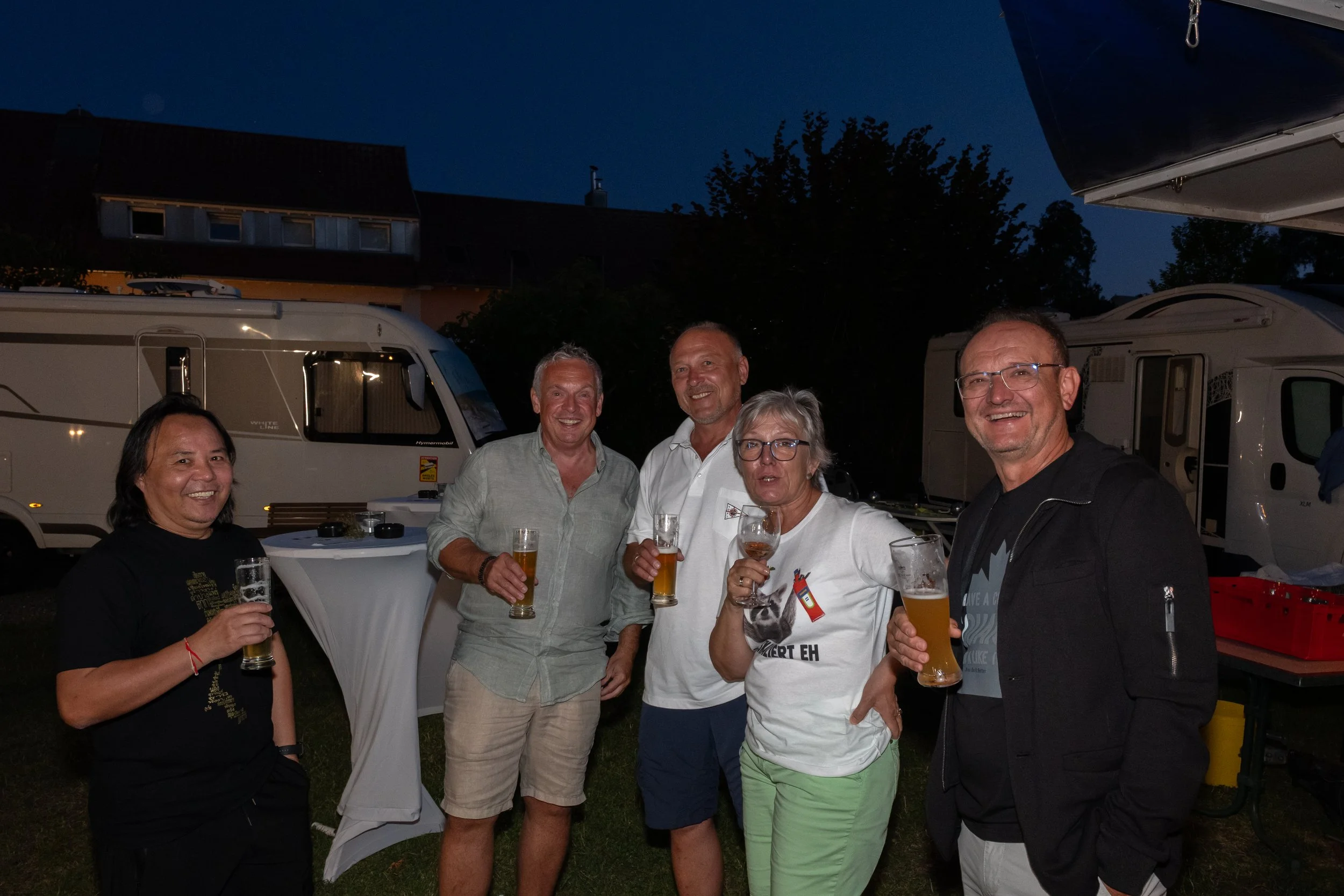 Eine Gruppe von sechs Menschen, die draußen abends Bier trinken, bei Zelten und Wohnmobilen im Hintergrund.