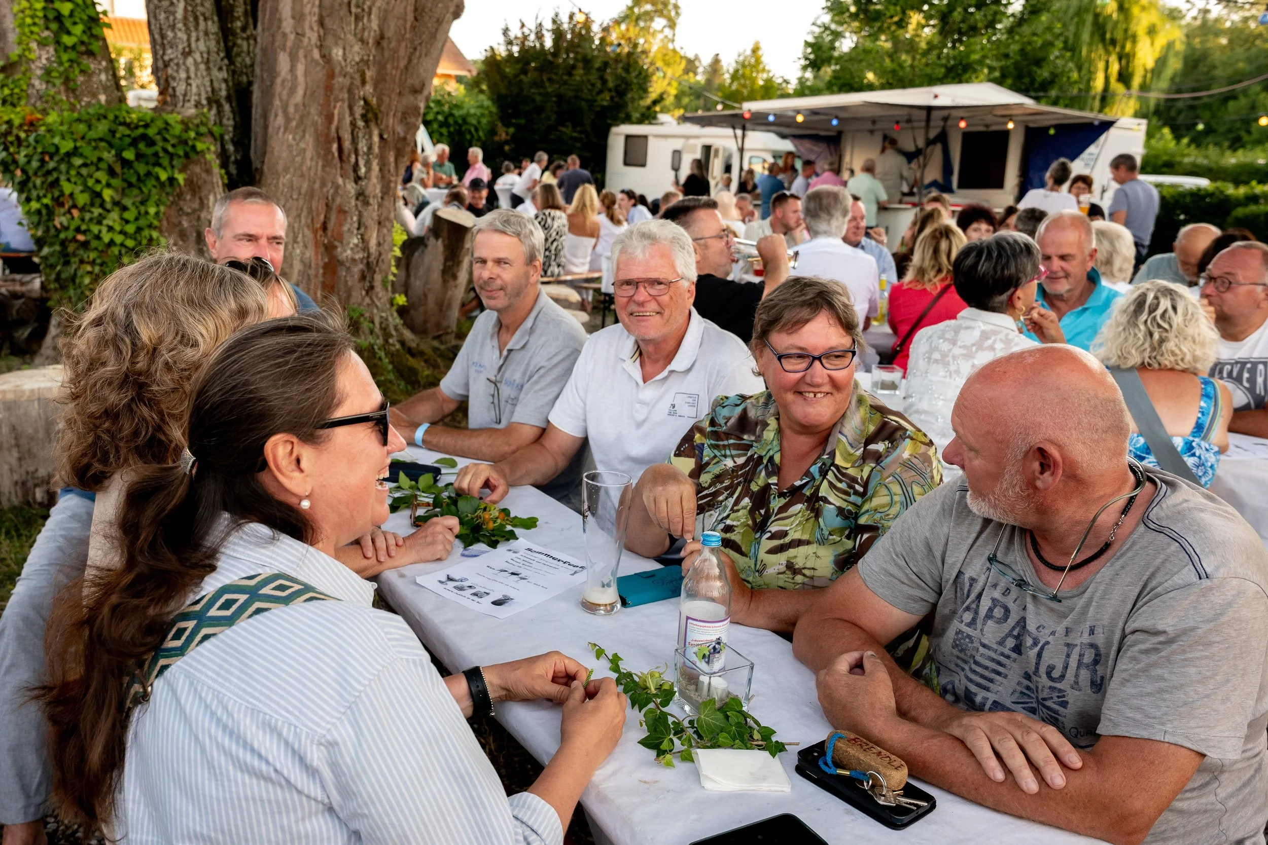 Menschen sitzen bei einem Outdoor-Fest an einem langen Tisch und unterhalten sich, im Hintergrund sind mehr Leute aufgestellt, unter Bäumen mit Lichterketten.