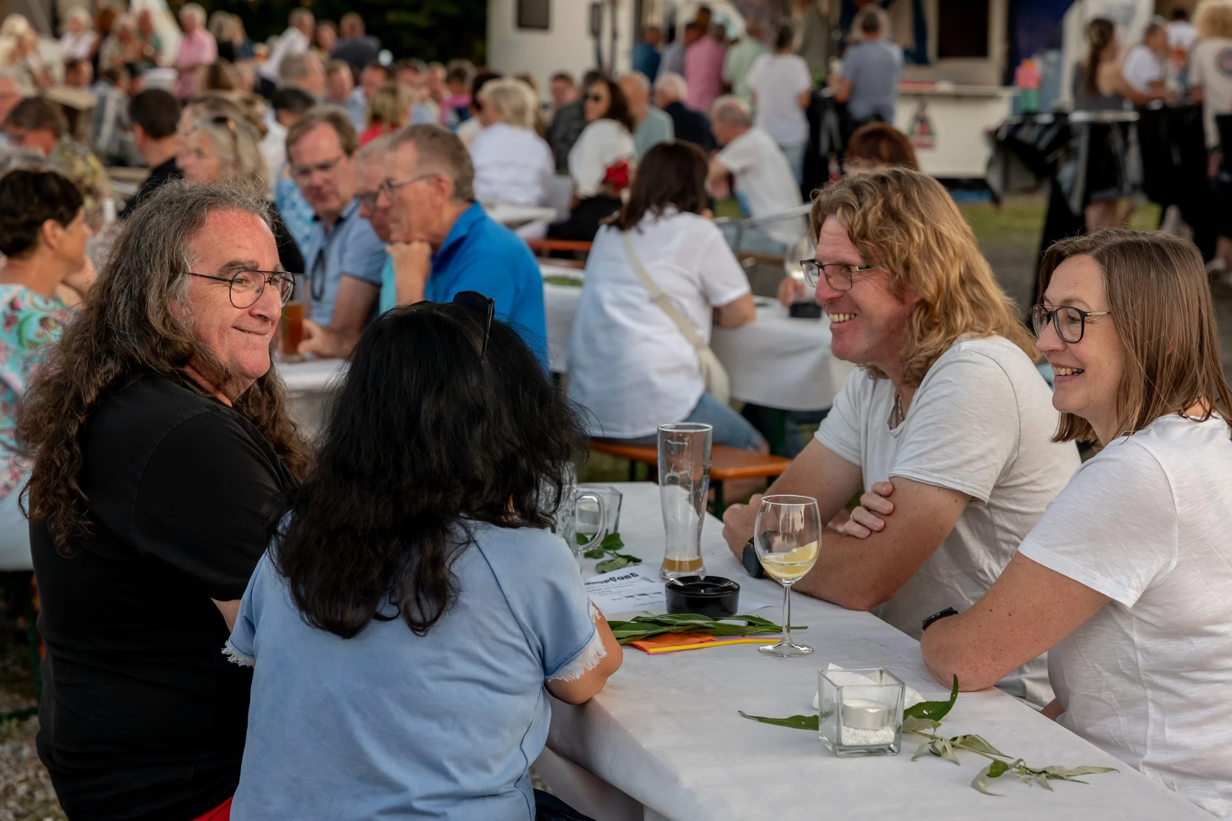 Menschen sitzen an einem langen Tisch bei einem Outdoor-Event, lachen und unterhalten sich.