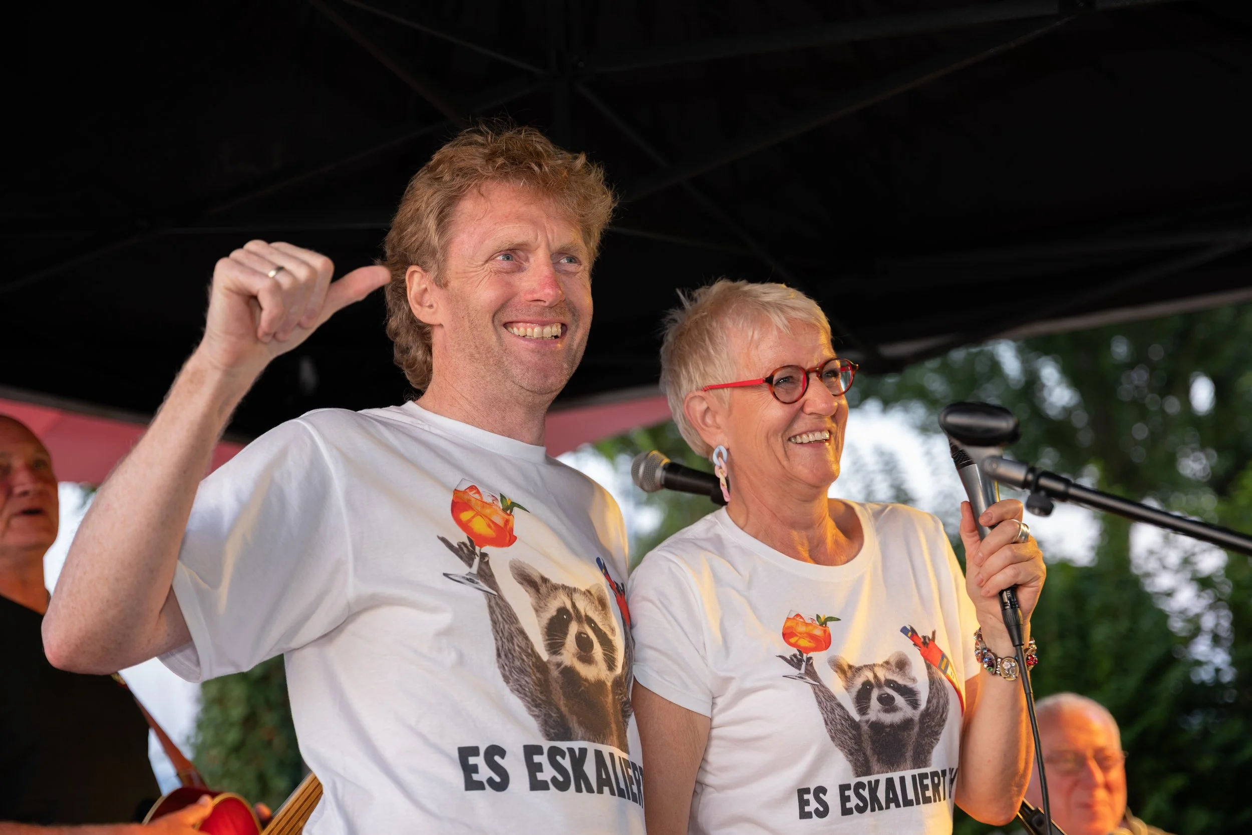 Zwei Menschen auf einer Bühne, die T-Shirts mit einem Waschbär und dem Text 'Es eskaliert' tragen, während sie lachen und eine Rede halten, im Hintergrund Musiker.