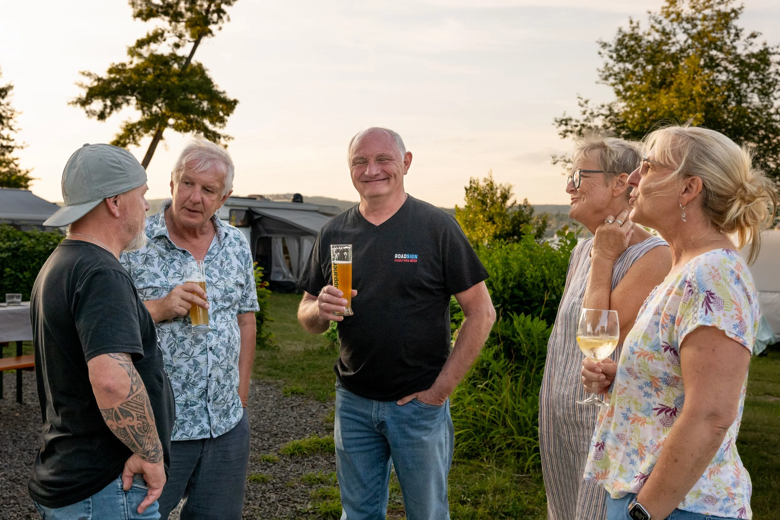 Fünf Erwachsene beim geselligen Beisammensein im Freien, bei Sonnenuntergang, mit Getränken in der Hand, lachend und im Gespräch.