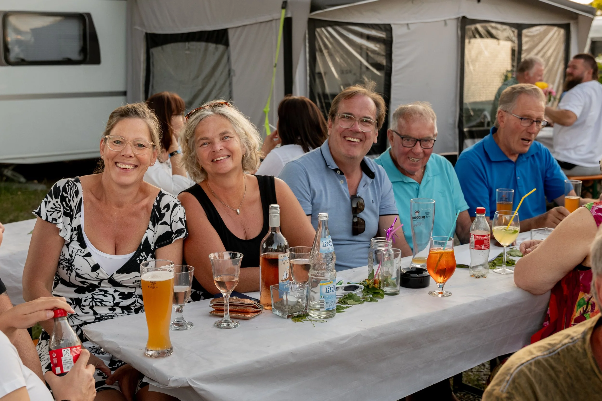 Menschen sitzen an einem langen Tisch im Freien, sie lachen und genießen Getränke bei einer Zusammenkunft.