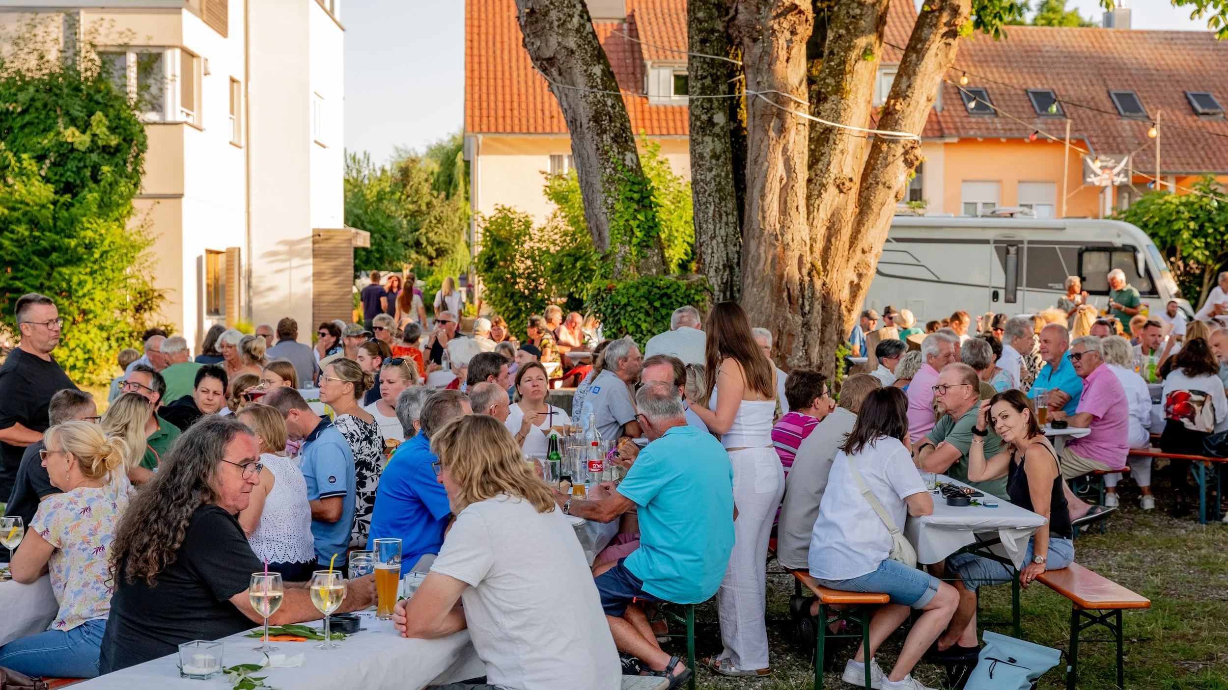 Menschen sitzen bei einem abendlichen Treffen im Freien unter Bäumen an langen Tischen, essen und trinken, während im Hintergrund ein Lastwagen parkt und mehr Leute zu sehen sind.