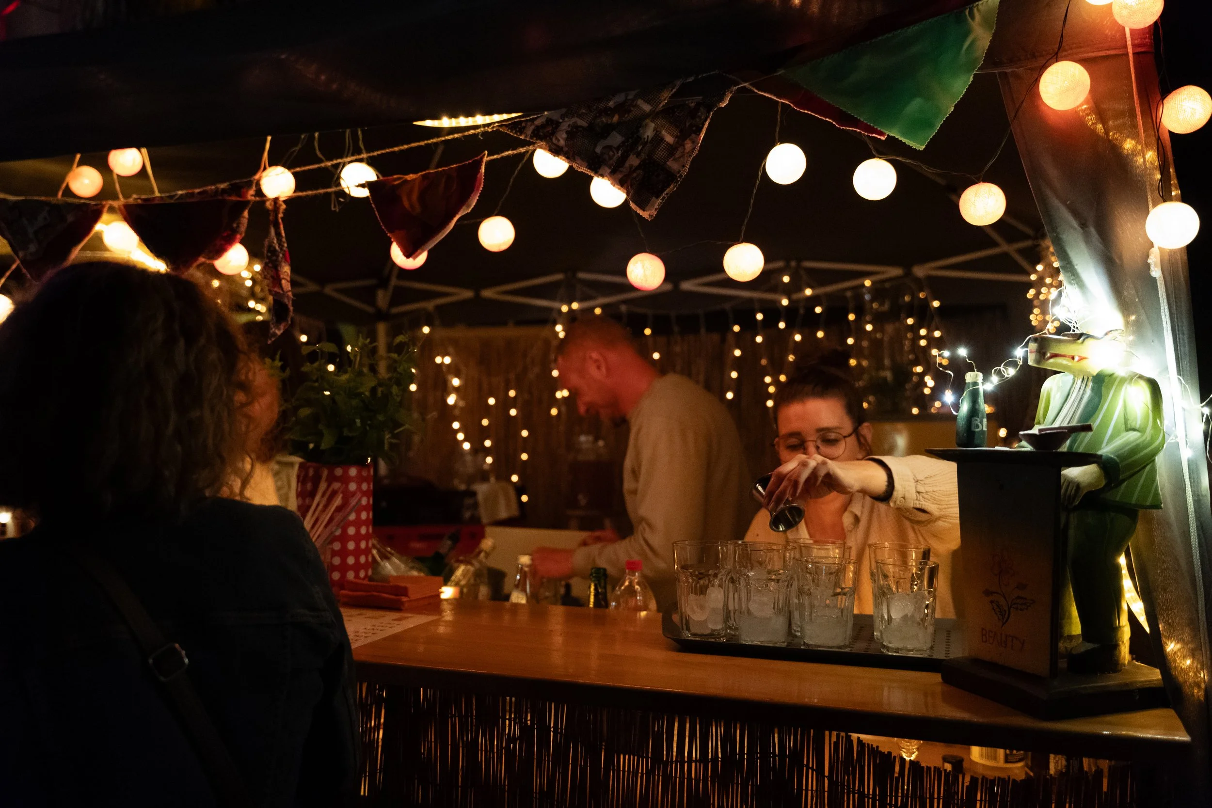 Blick auf eine gemütliche Bar bei Nacht, beleuchtet mit bunten Lichterketten, mit einem Barkeeper, der Getränke zubereitet, und Gästen, die an der Bar sitzen und Getränke genießen.