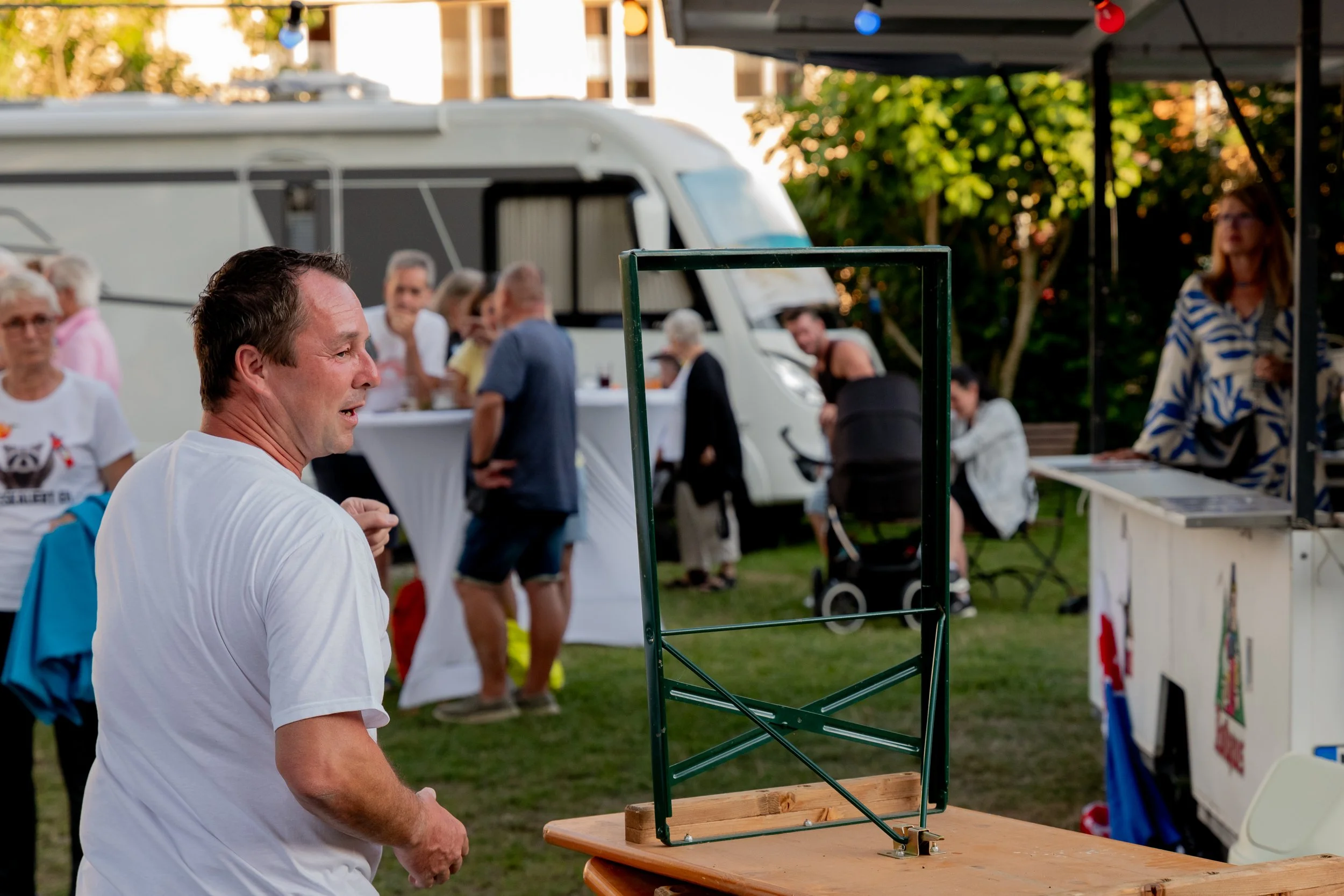 Ein Mann bei einem Outdoor-Festwerk mit einer aufgestellten Tischtennisplatte, im Hintergrund sind mehrere Menschen rund um Stehtische, ein Wohnmobil und ein Stand mit Frau.