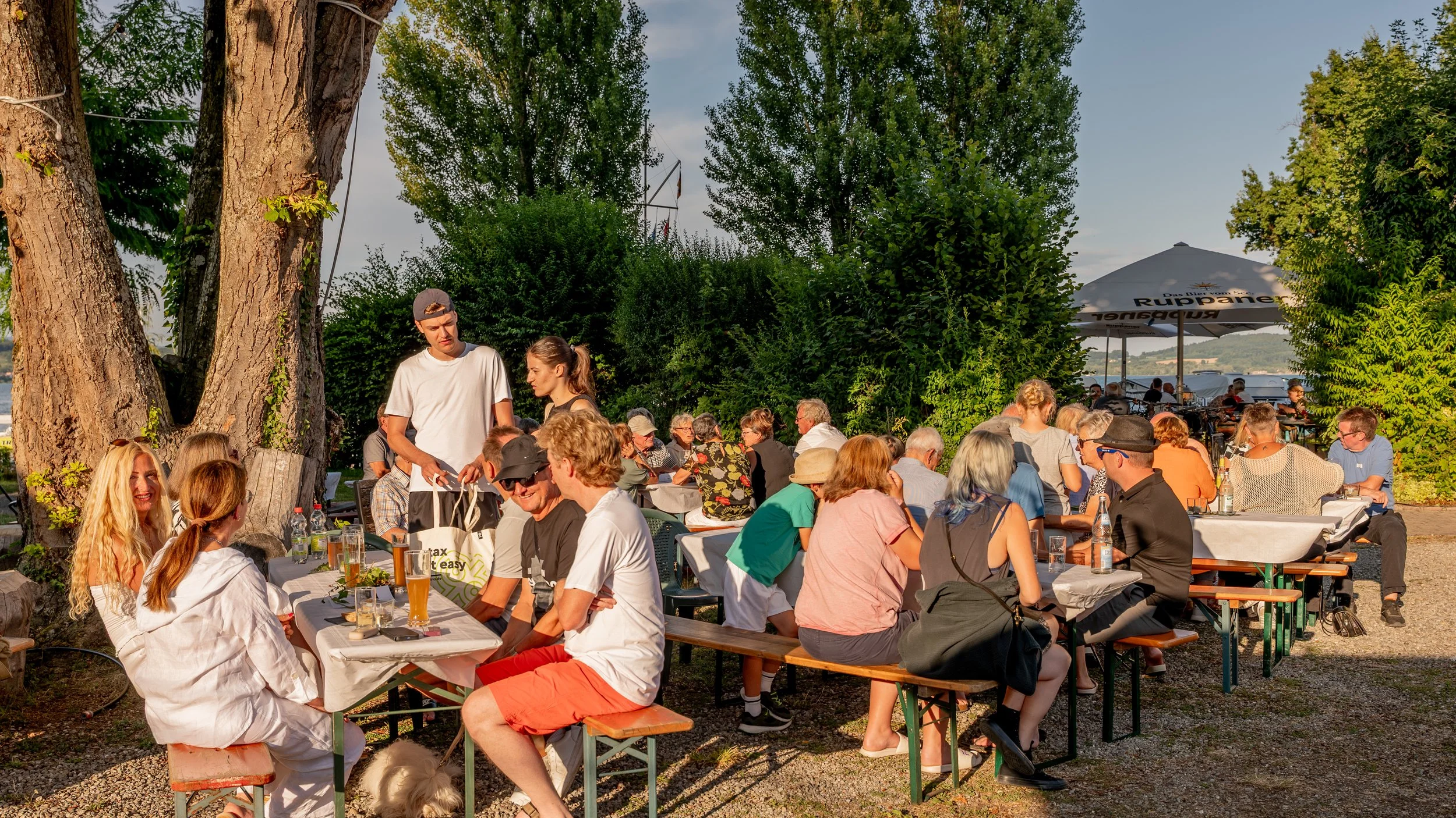 Menschen sitzen bei einem Tageslichtabend im Freien an Tischen unter Bäumen. Ein großer Sonnenschirm ist zu sehen, im Hintergrund sind weitere Gäste und Bäume. Es sieht nach einer geselligen Atmosphäre aus.