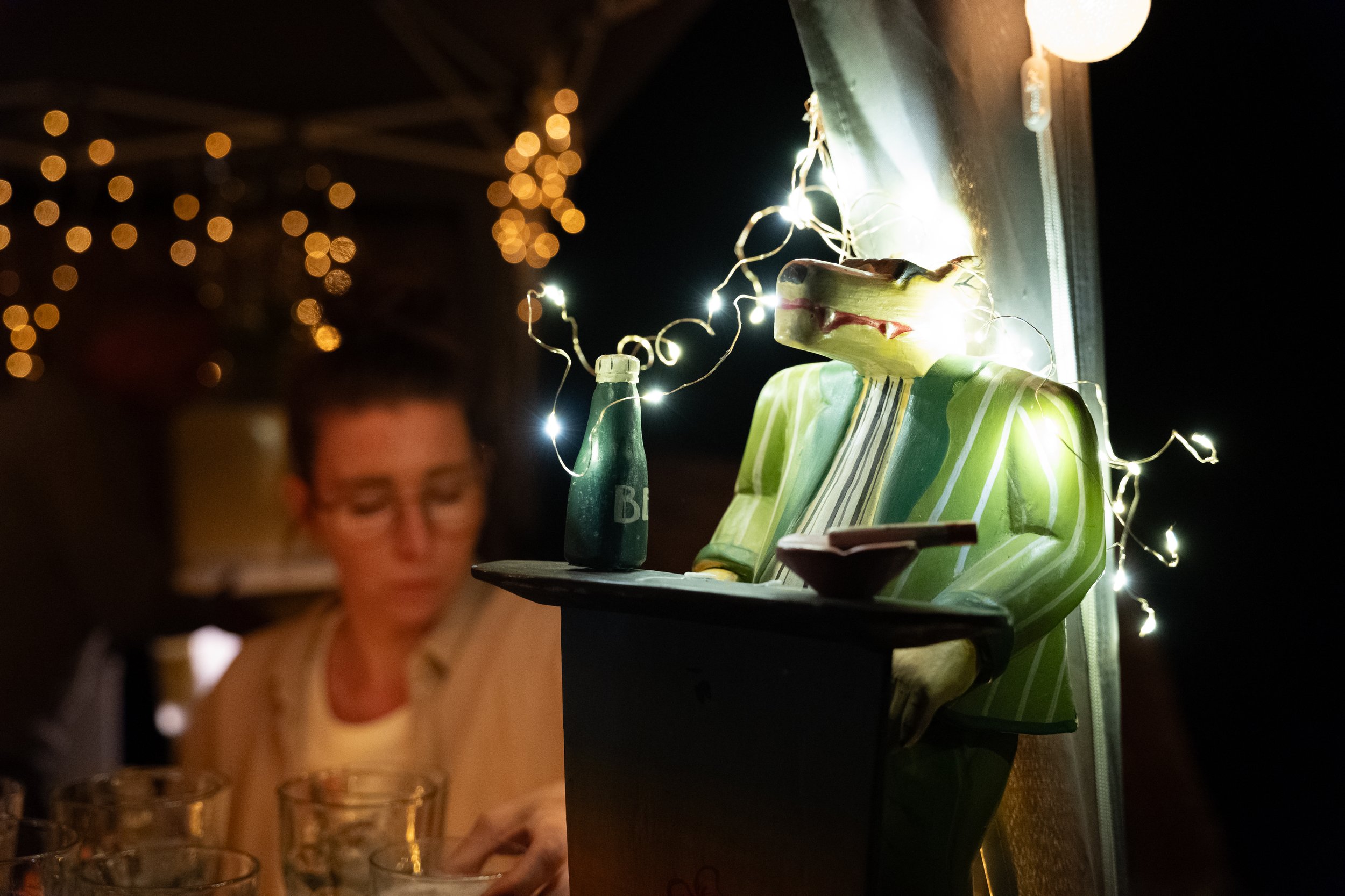Ein bunter, vorgesetzter Drachenkopf mit grün-weiß-gestreiftem Hemd, der auf einer Theke sitzt, umgeben von Weihnachtslichtern, im Hintergrund eine Frau mit Brille, die Getränke einschenkt, bei Nacht.