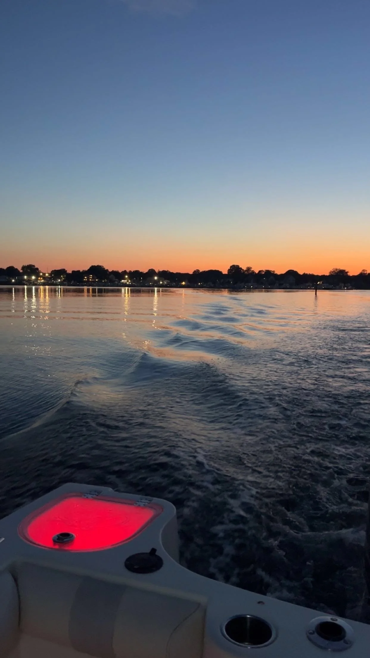 View of a lake at sunset with a boat's sonar or depth finder display in the foreground, showing a red glow, and houses along the shoreline with lights reflecting on the water.