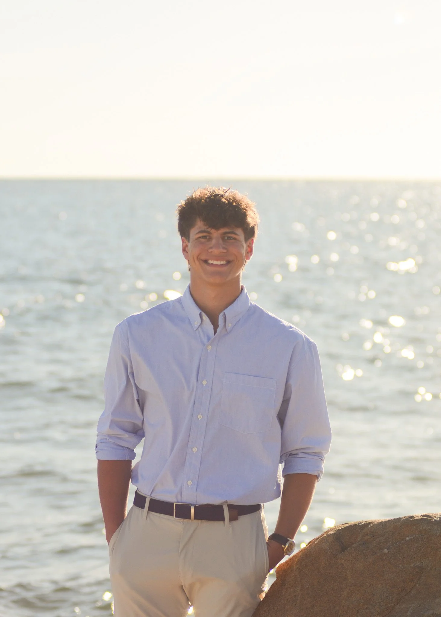 A young man in a white button-up shirt and beige pants standing outdoors near water, smiling at the camera during sunset or sunrise.