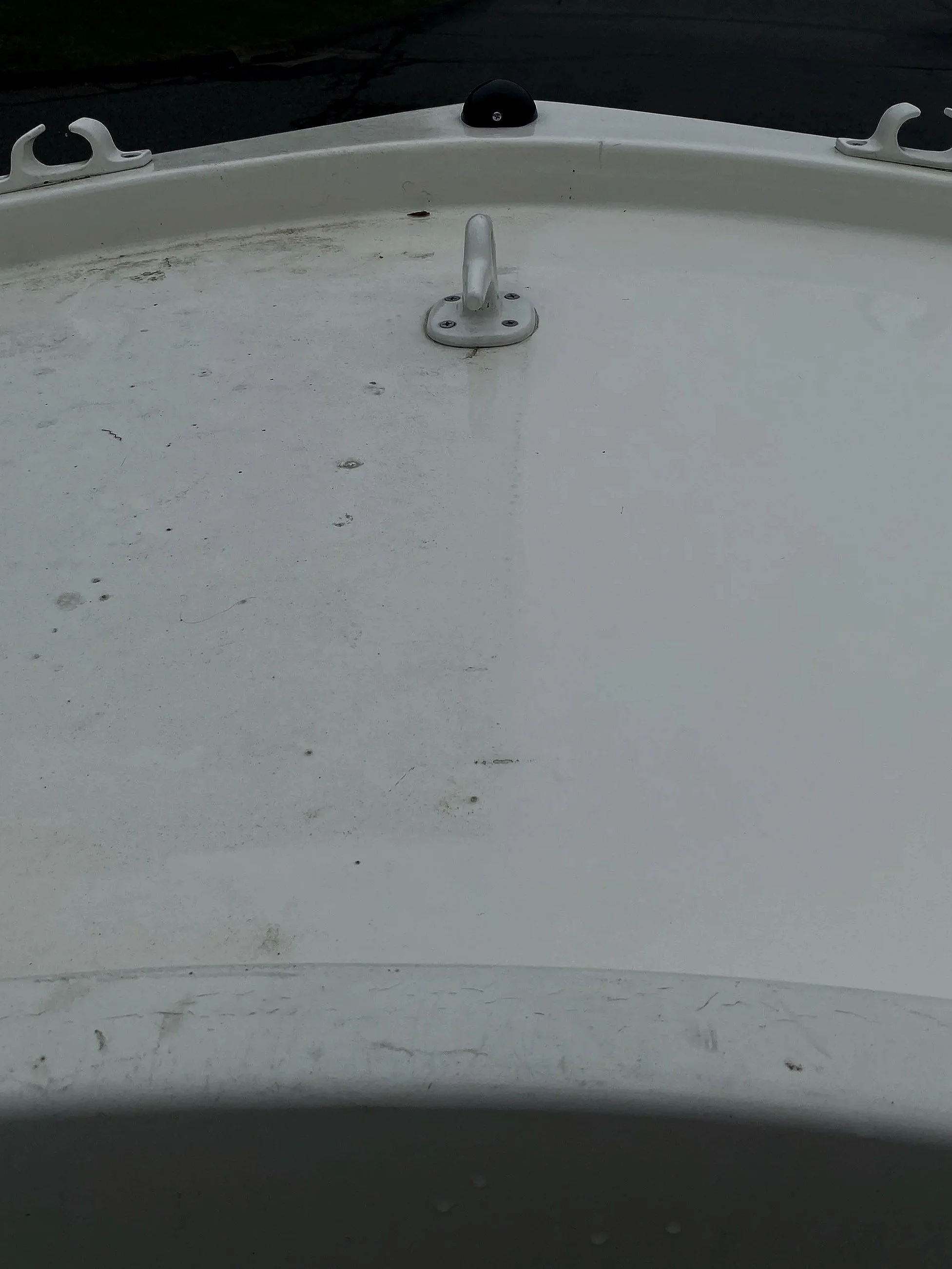 The top of a white boat with a metal cleat, a metal mount for a flagpole, and a black antenna or sensor. There are some dirt and scratches on the surface of the boat. The background shows a dark water surface.