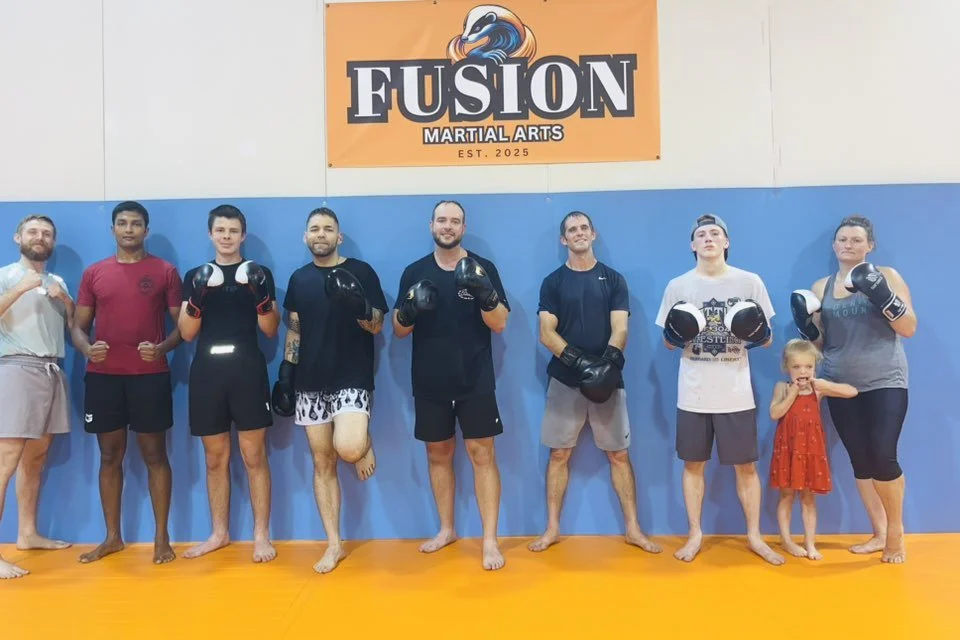 Group of nine people, including children, standing inside a martial arts gym with a sign that reads "FUSION MARTIAL ARTS" on the wall. Some are wearing boxing gloves, and the setting has a blue wall and orange floor.