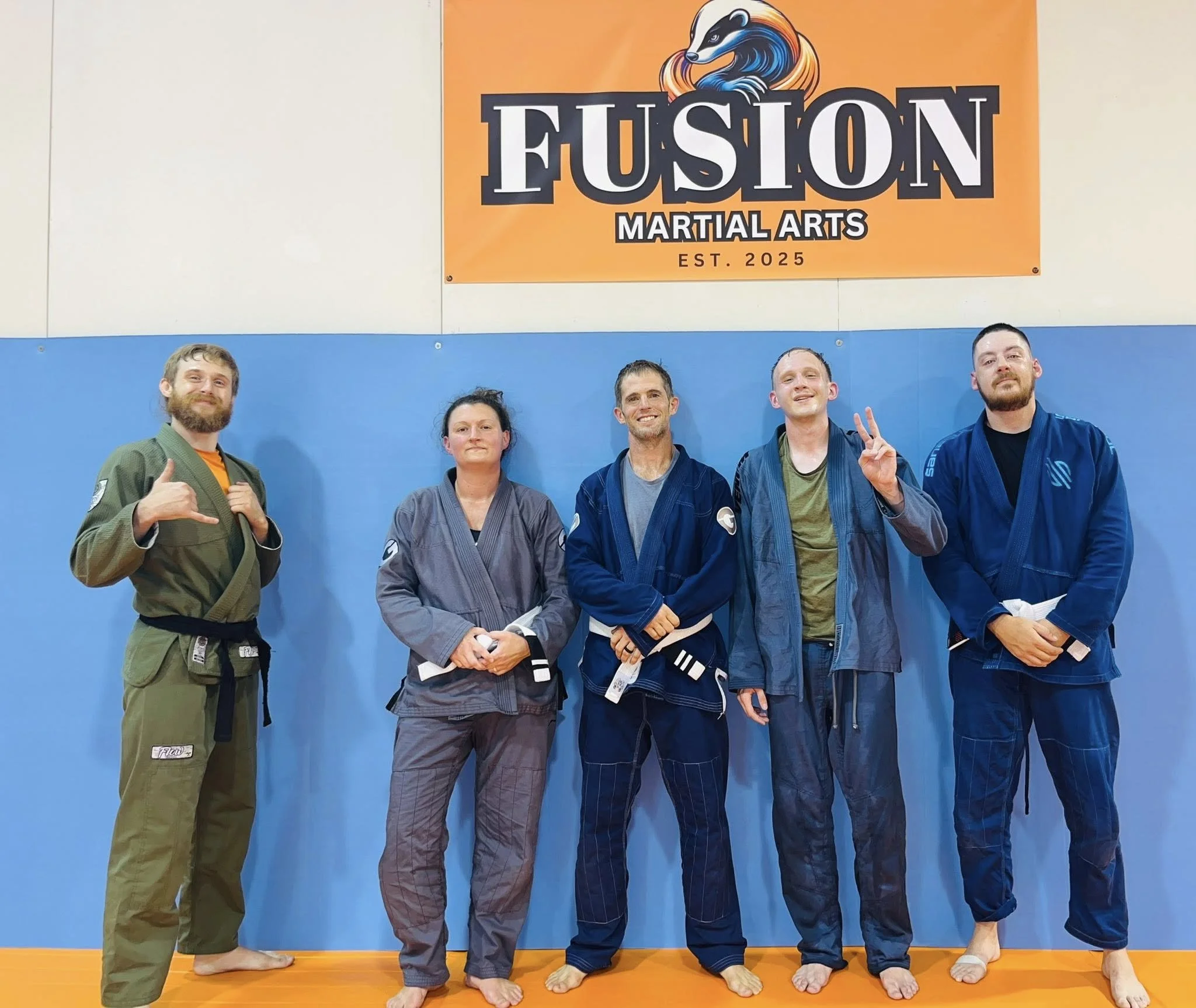 Group of five people dressed in Brazilian Jiu-Jitsu gis standing on a blue mat in a martial arts gym, with a Fusion Martial Arts banner in the background.