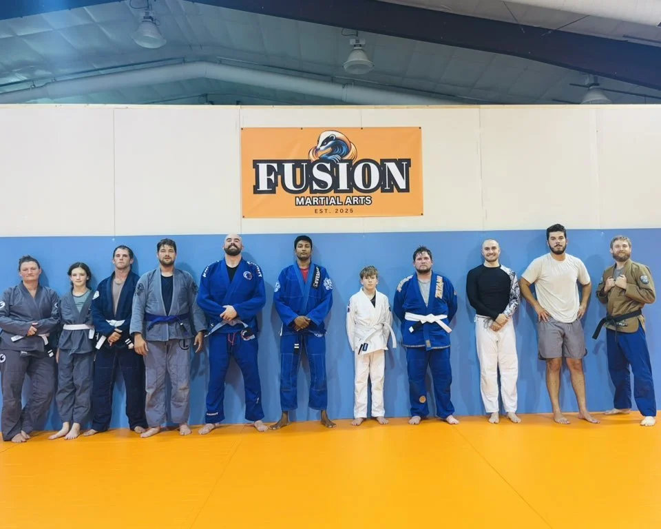 Group of martial artists standing in a row in a gym, wearing gis and belts, with a 'Fusion Martial Arts' sign on the wall behind them.