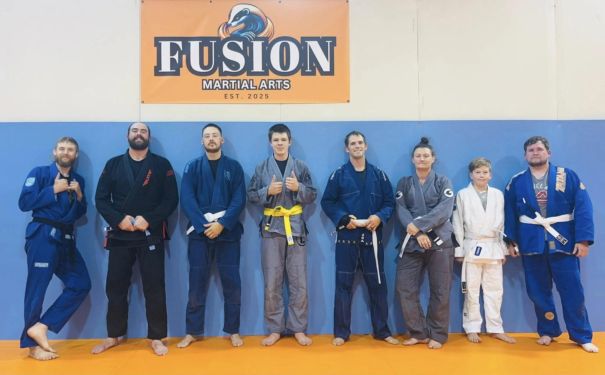 A group of nine martial artists in jiu-jitsu gi uniforms standing in a line on an orange mat, posing for a photo with a Fusion Martial Arts banner above them.
