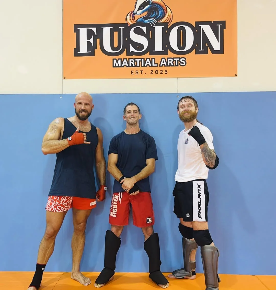 Three men in martial arts gear standing in a gym with a banner that says Fusion Martial Arts. The man on the left wears red shorts, a black tank top, red gloves, and no shoes. The man in the middle wears red shorts, a dark t-shirt, and black protecti