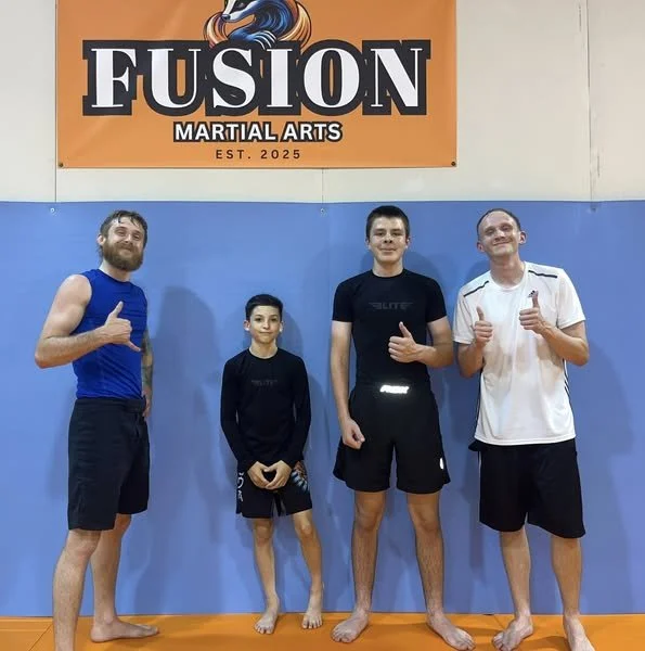 Four males standing on a blue mat at a martial arts gym, in front of an orange banner that says 'Fusion Martial Arts EST. 2025', giving thumbs up.