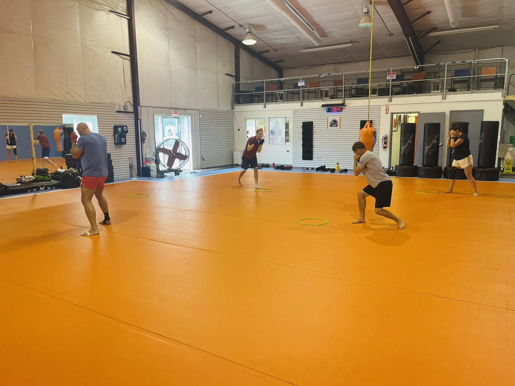 People practicing boxing drills in a gym with orange mats, punching bags, and a second-floor balcony.