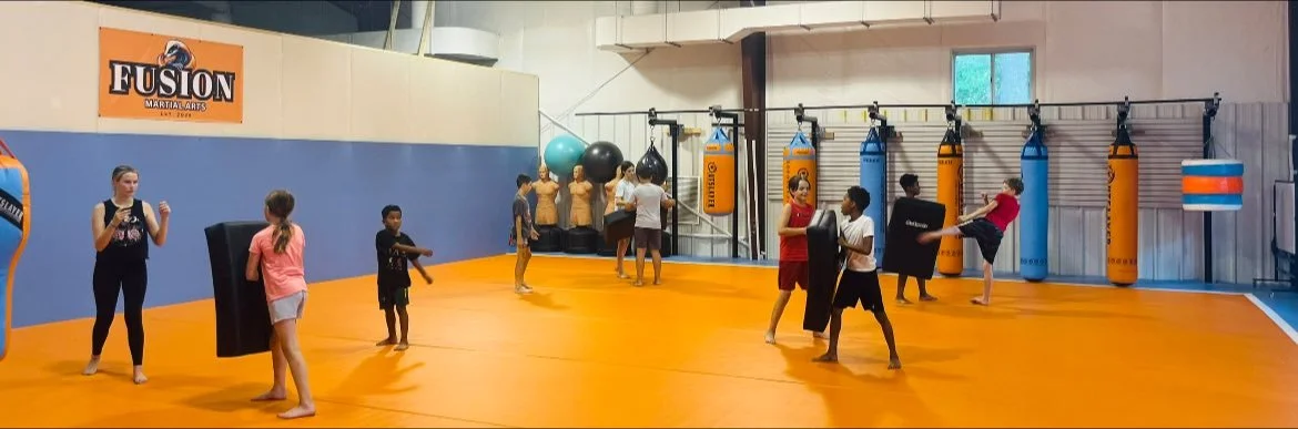 Children and adults practicing martial arts with punching pads in a gym with orange mat flooring and punching bags.
